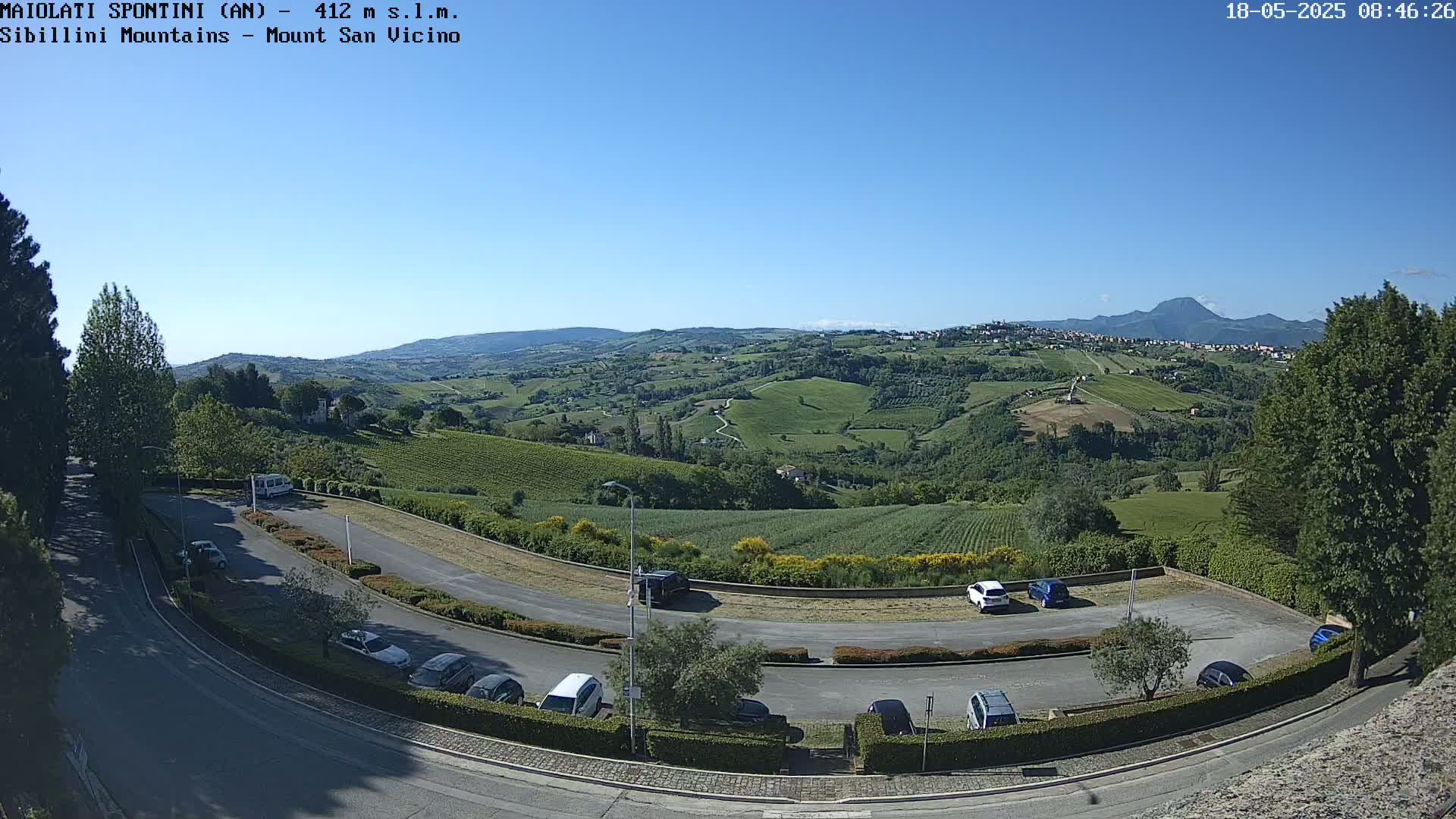 A parking lot with several cars curves around a hillside overlooking a sunny valley of vineyards and distant mountains under a clear blue sky.