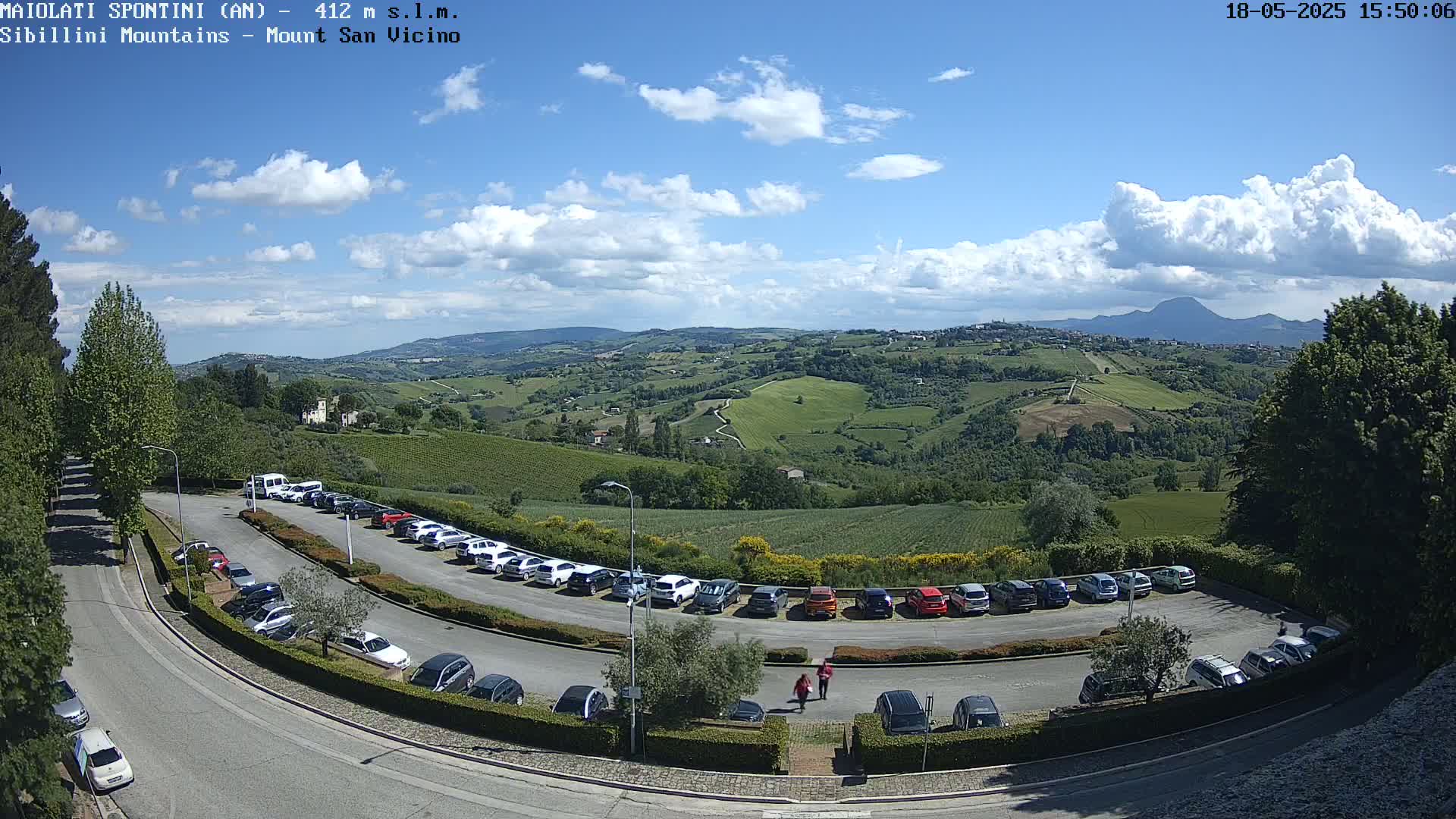 A partly cloudy blue sky overlooks a hillside parking lot full of cars and a sprawling green valley dotted with trees and buildings, extending to distant mountains.