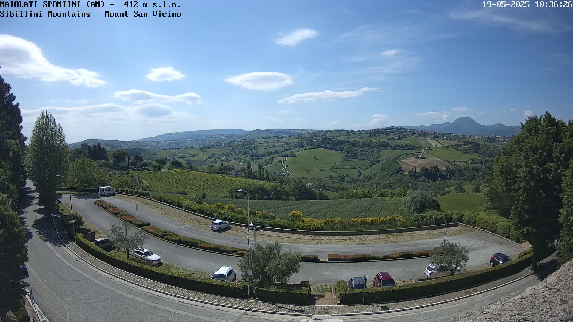 A mostly sunny day reveals a hilly landscape with vineyards and a few cars parked in a curved lot next to a road.