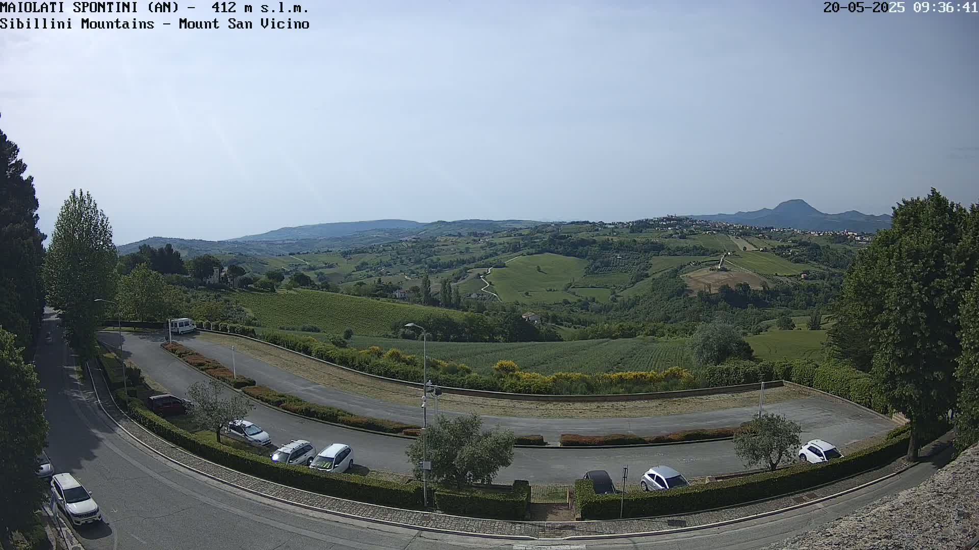 A parking lot with several cars overlooks a sunlit valley of rolling green hills and vineyards under a clear sky.