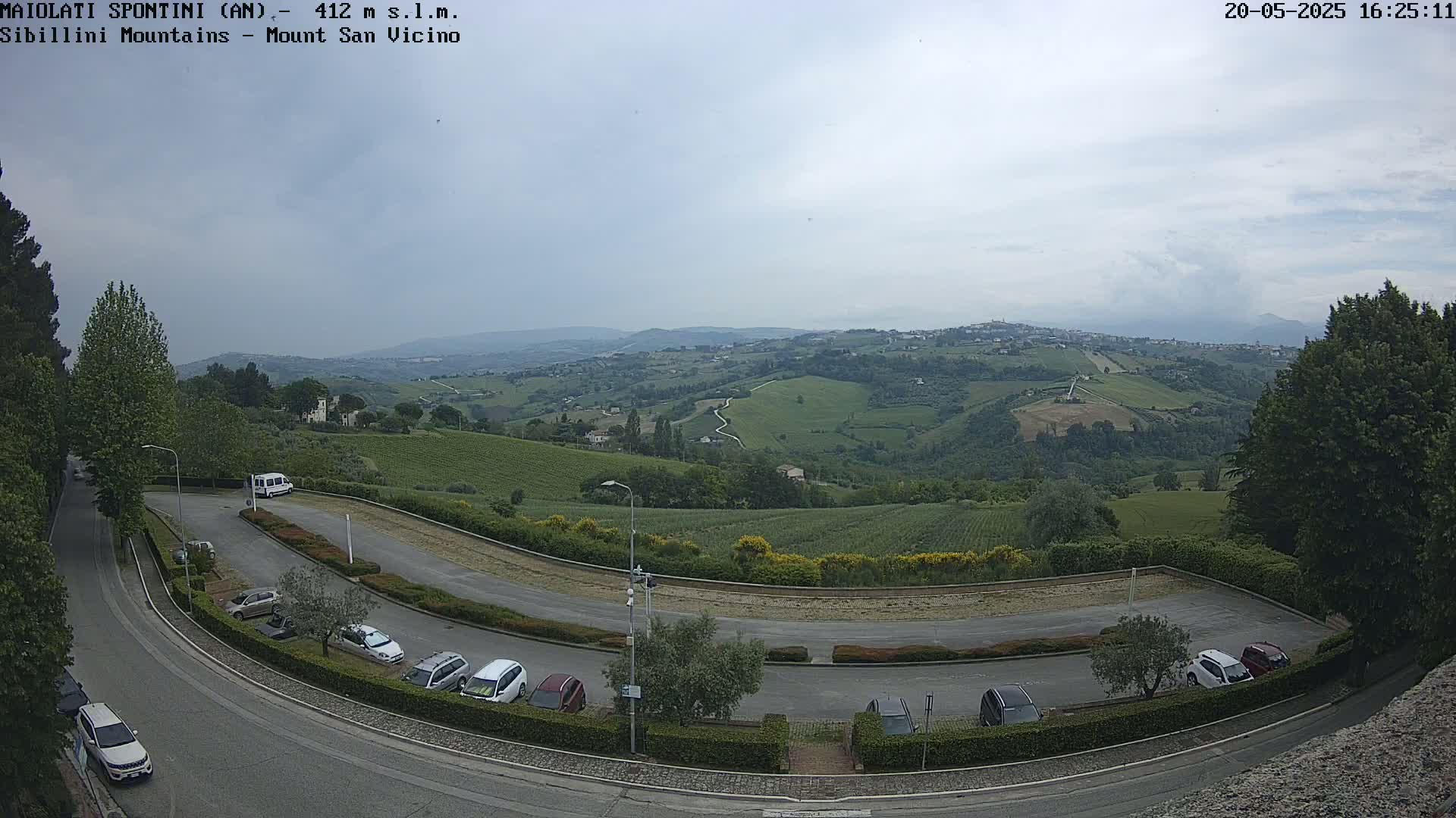A mostly cloudy outdoor scene shows a curved parking lot with several cars, overlooking a hilly landscape of vineyards and farmlands under an overcast sky.