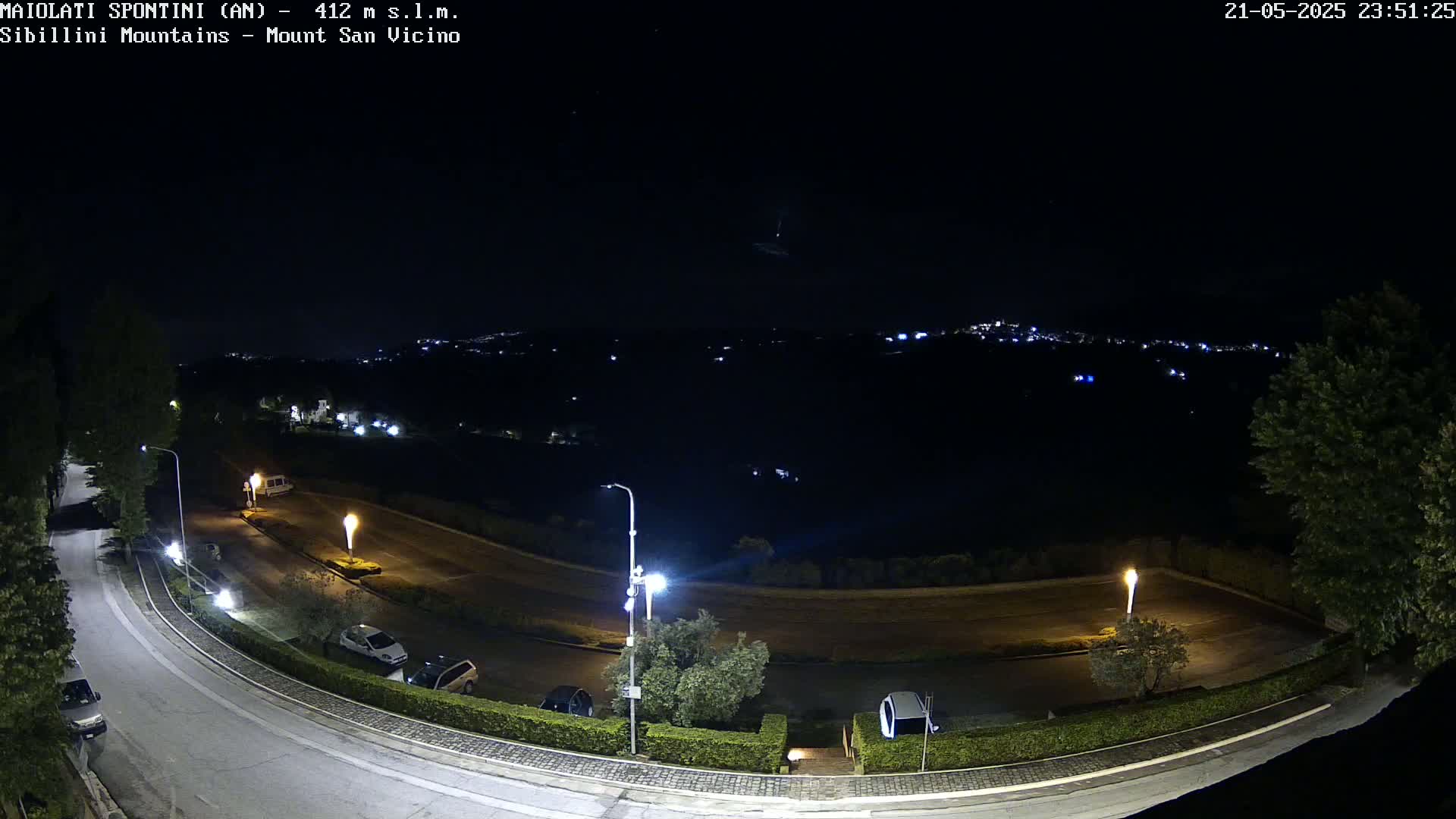 A nighttime, high-angle view of a curving road with parked cars, illuminated by streetlights, overlooking a dark valley with distant city lights.