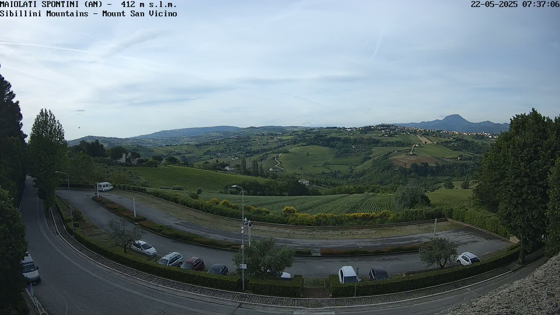 A mostly sunny day overlooking a hilly landscape with vineyards and a parking lot containing several cars, viewed from an elevated vantage point.