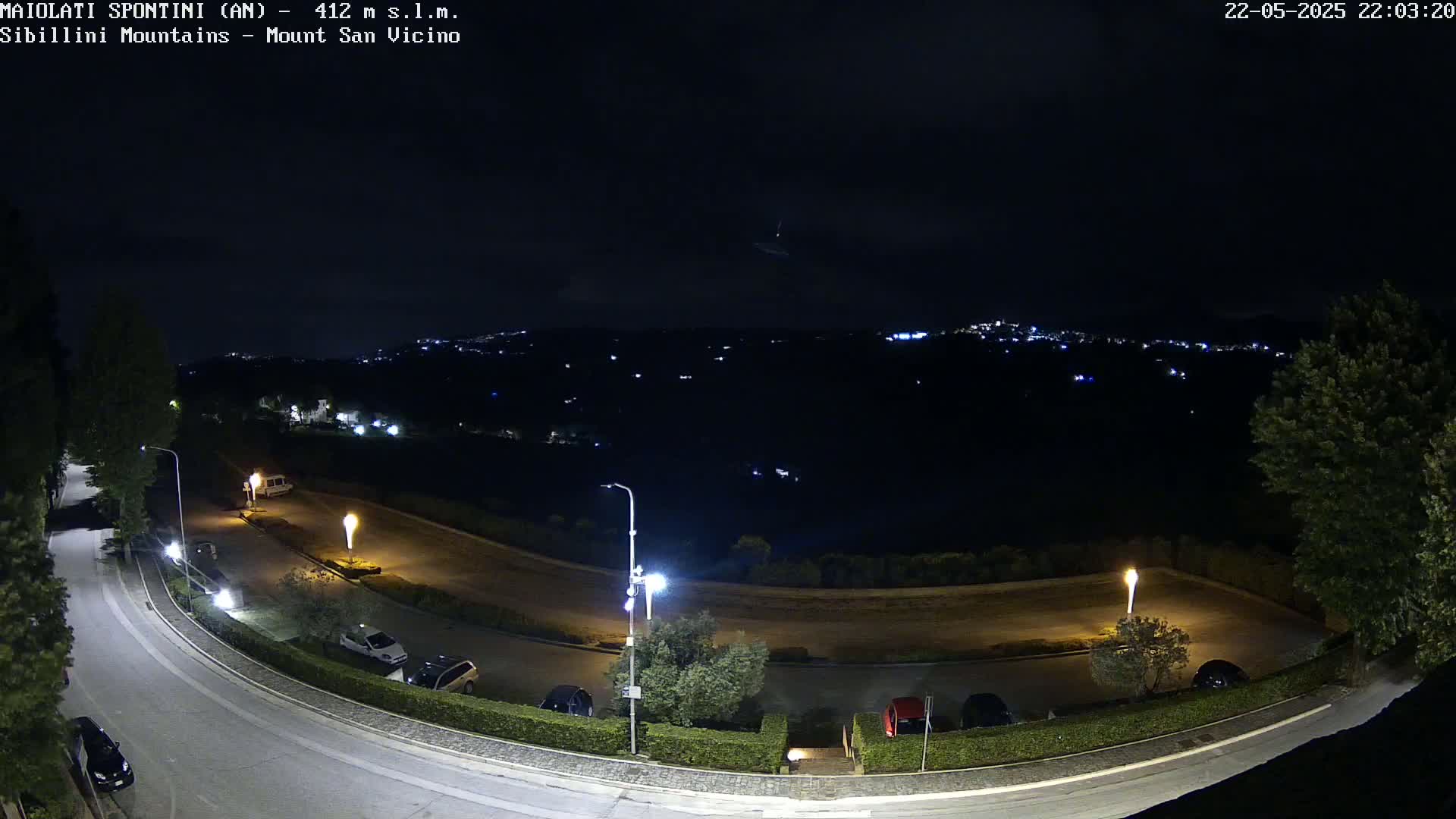 A nighttime, overhead view of a parking lot and curving road, illuminated by streetlights, overlooks a dark, distant town nestled in a valley under a clear night sky.