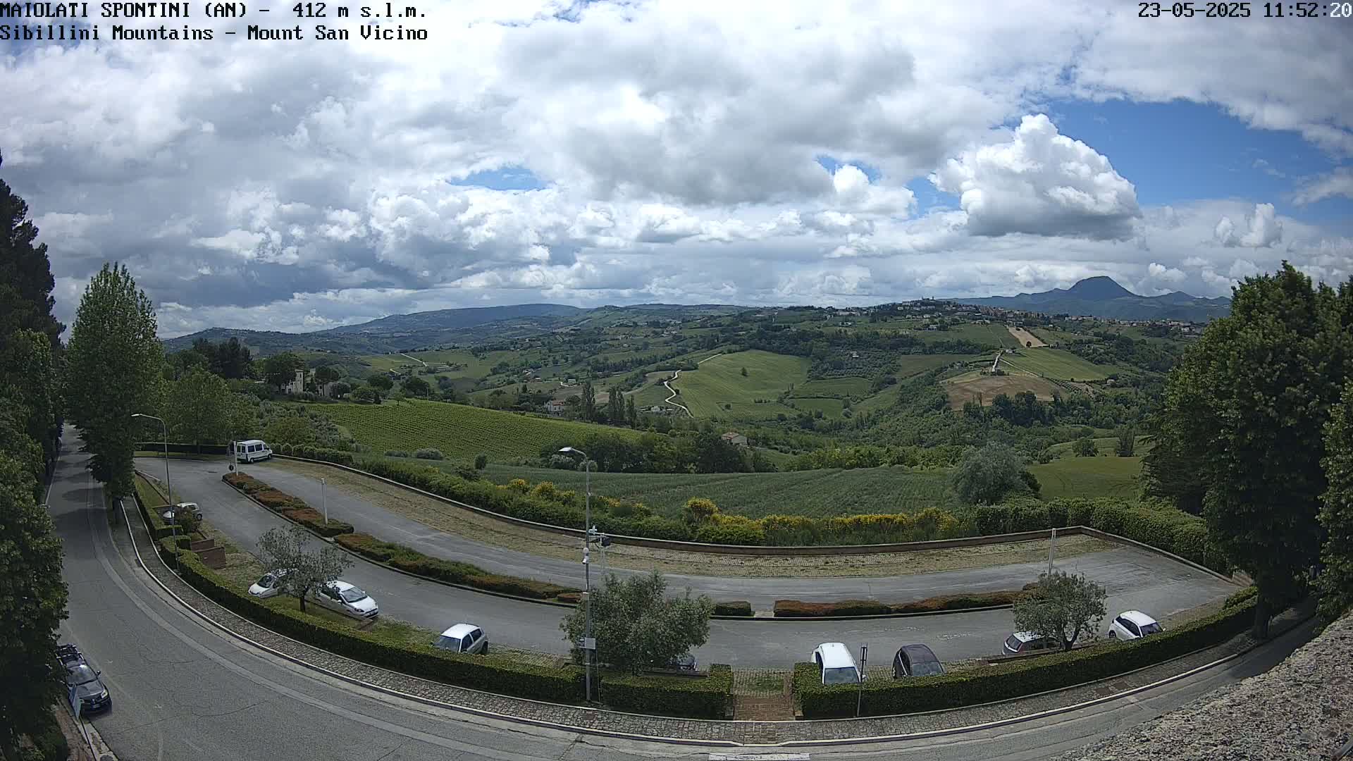 A partly cloudy day reveals a sweeping view of rolling green hills and a parking lot with several cars, overlooking a town nestled in the valley below.