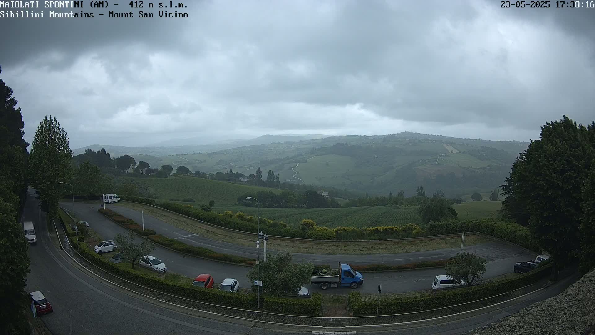 An overcast sky overlooks a hilly landscape with parked cars in a curved lot, and a road leading to a view of rolling green hills.