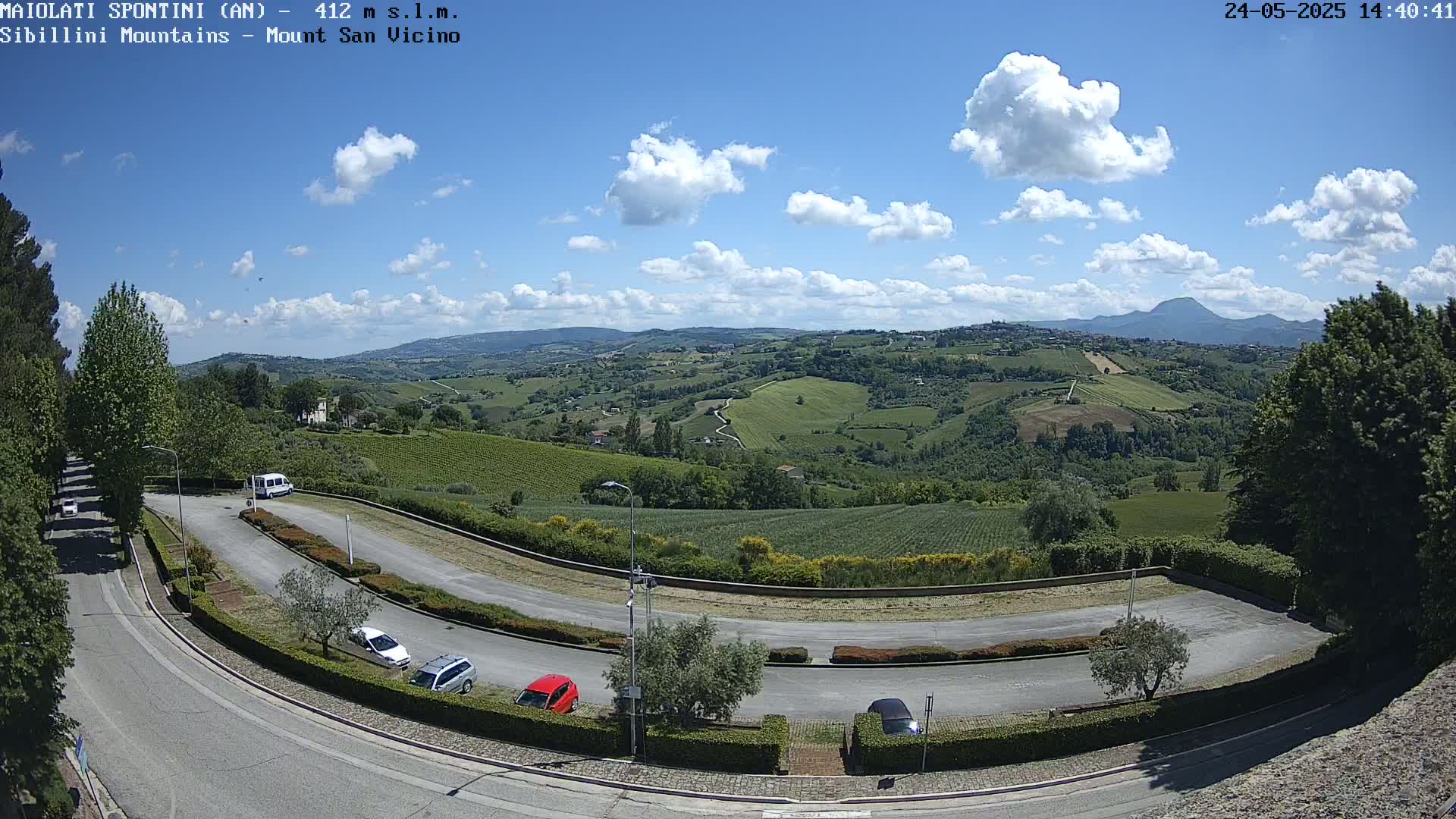 A winding road with parked cars curves through a parking lot overlooking a sunny, partly cloudy landscape of rolling green hills and distant mountains.