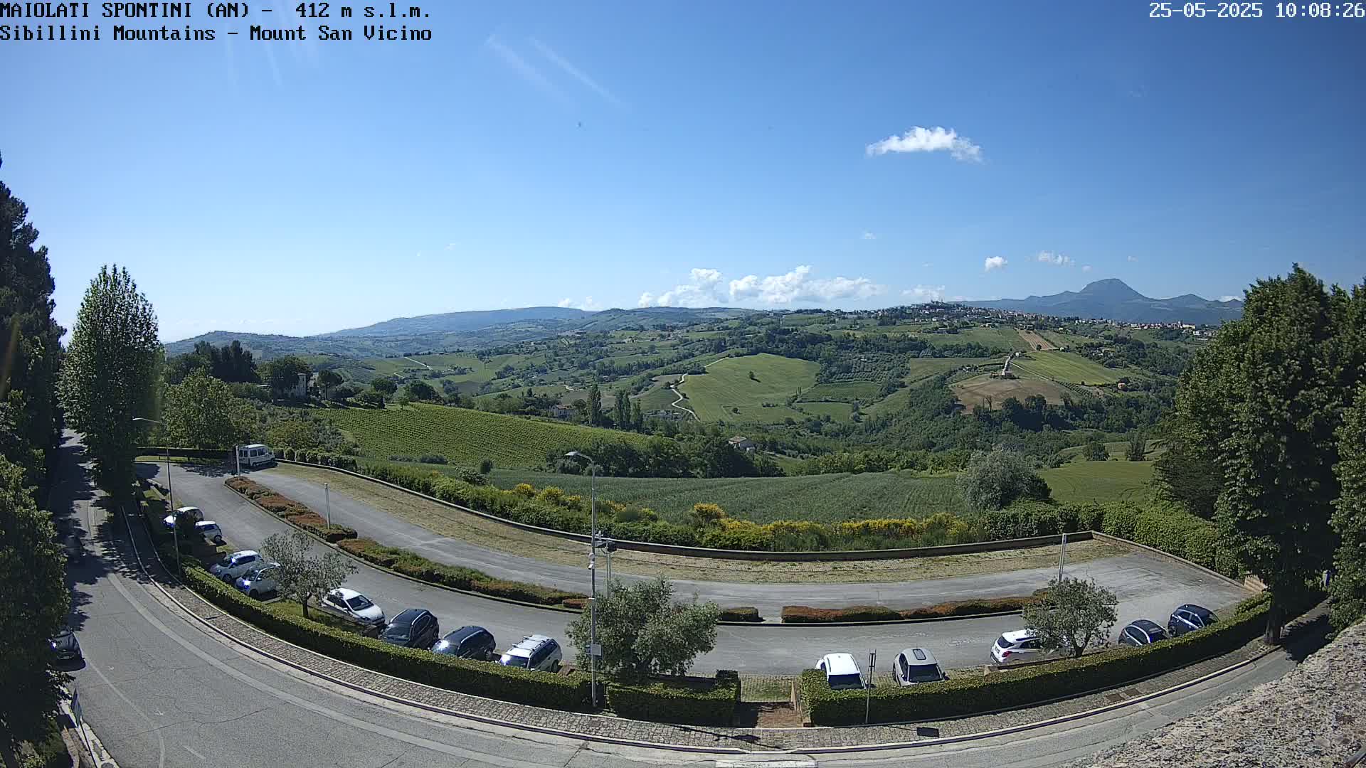 A mostly sunny outdoor view of a parking lot overlooking a hilly landscape with green fields and vineyards under a clear blue sky.