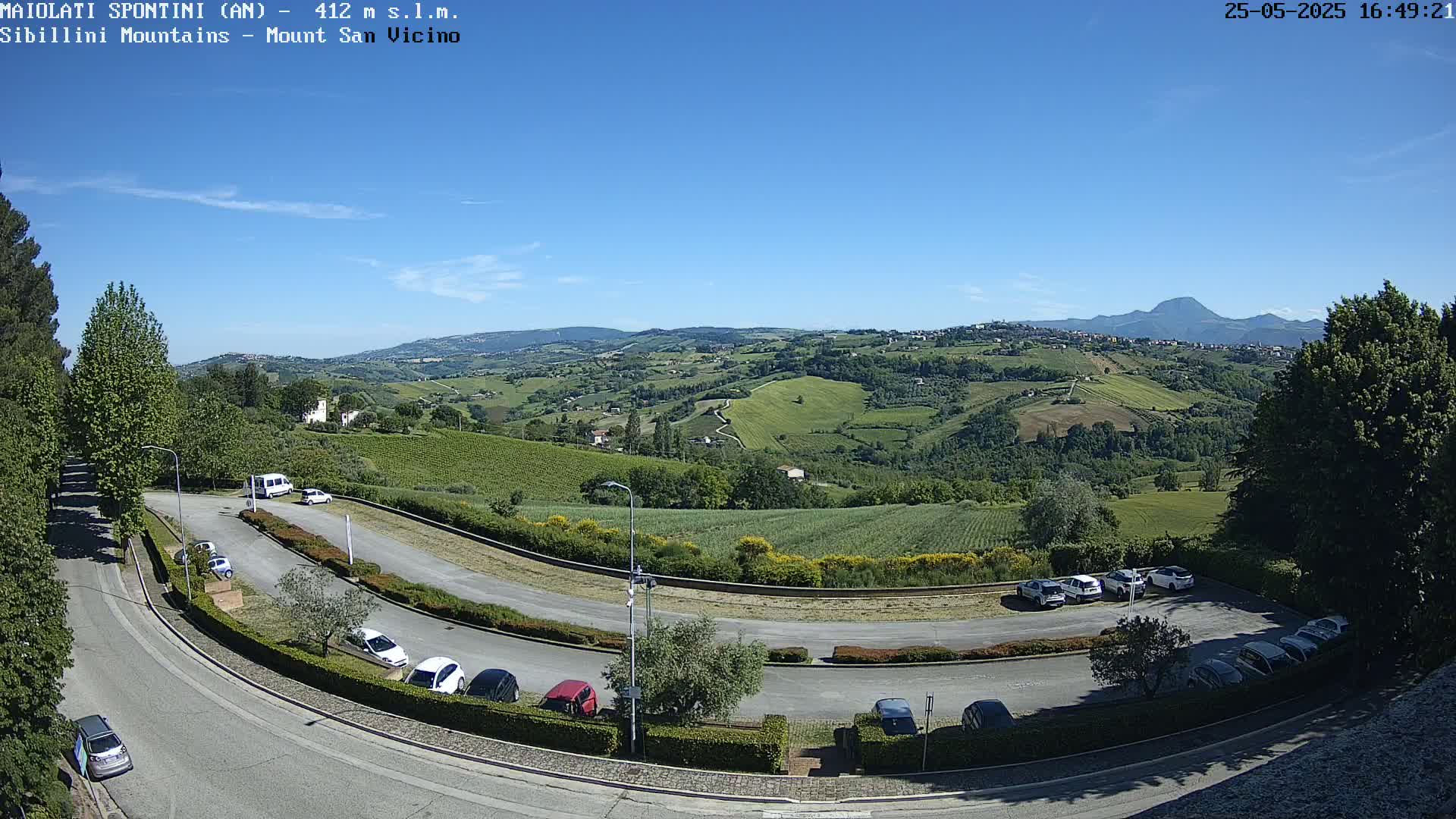 A sunny, clear day reveals a panoramic view of rolling green hills, vineyards, and a parking lot with several cars, overlooked by a road curving through the landscape.
