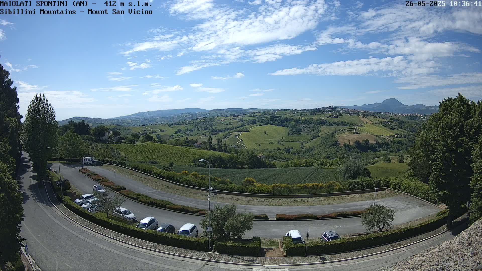 A parking lot with several cars is situated on a hill overlooking a vast, verdant landscape under a mostly sunny sky.