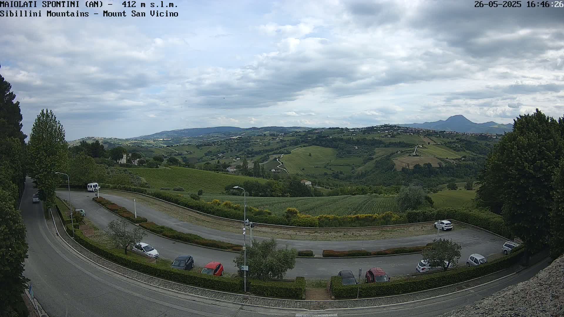 A parking lot with several cars overlooks a gently rolling landscape of green hills and vineyards under a mostly cloudy sky.