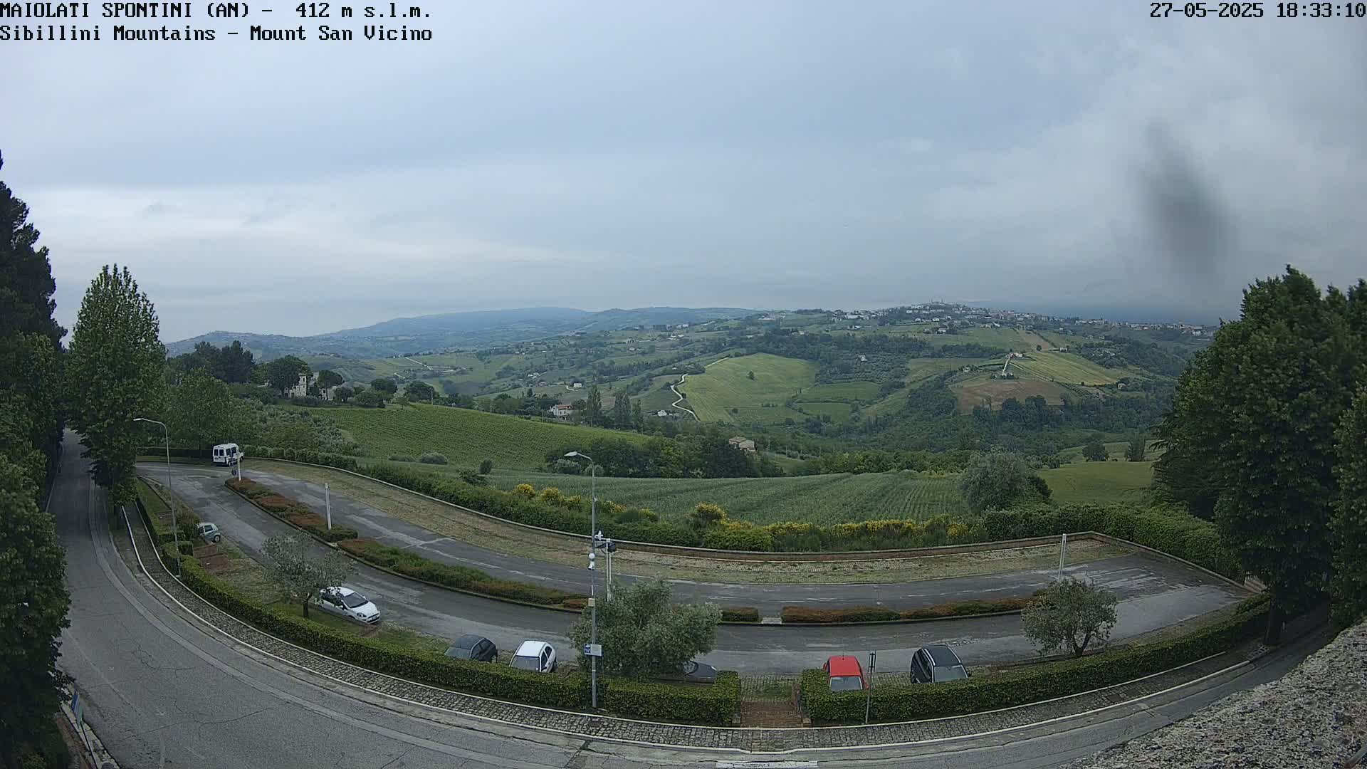 A winding road curves through a parking lot overlooking a hilly landscape of green fields and distant buildings under an overcast sky.