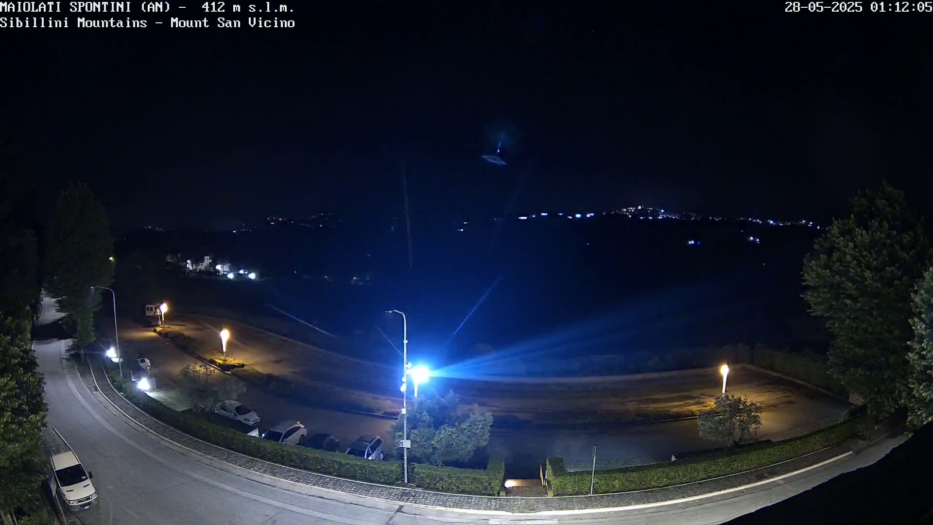 A nighttime, high-angle view shows a parking lot and curved road illuminated by streetlights, with a distant town visible in the background under a clear, dark sky.