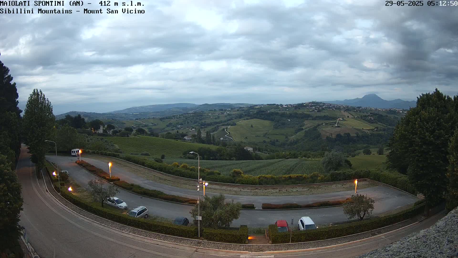 A winding road with parked cars curves through a hilly landscape under a cloudy sky, overlooking a valley with distant mountains.