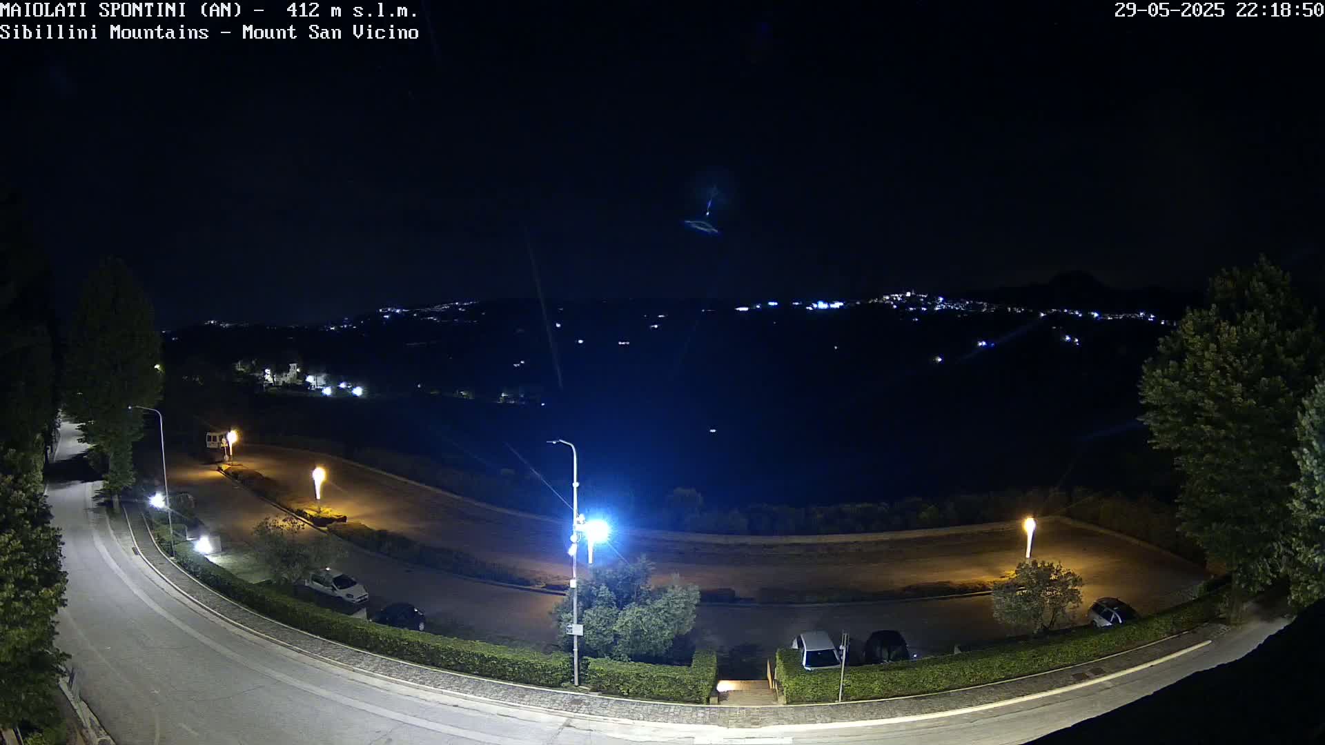 A nighttime, high-angle view of a curving road with parked cars and streetlights, overlooking a distant, dimly lit town nestled in dark hills.