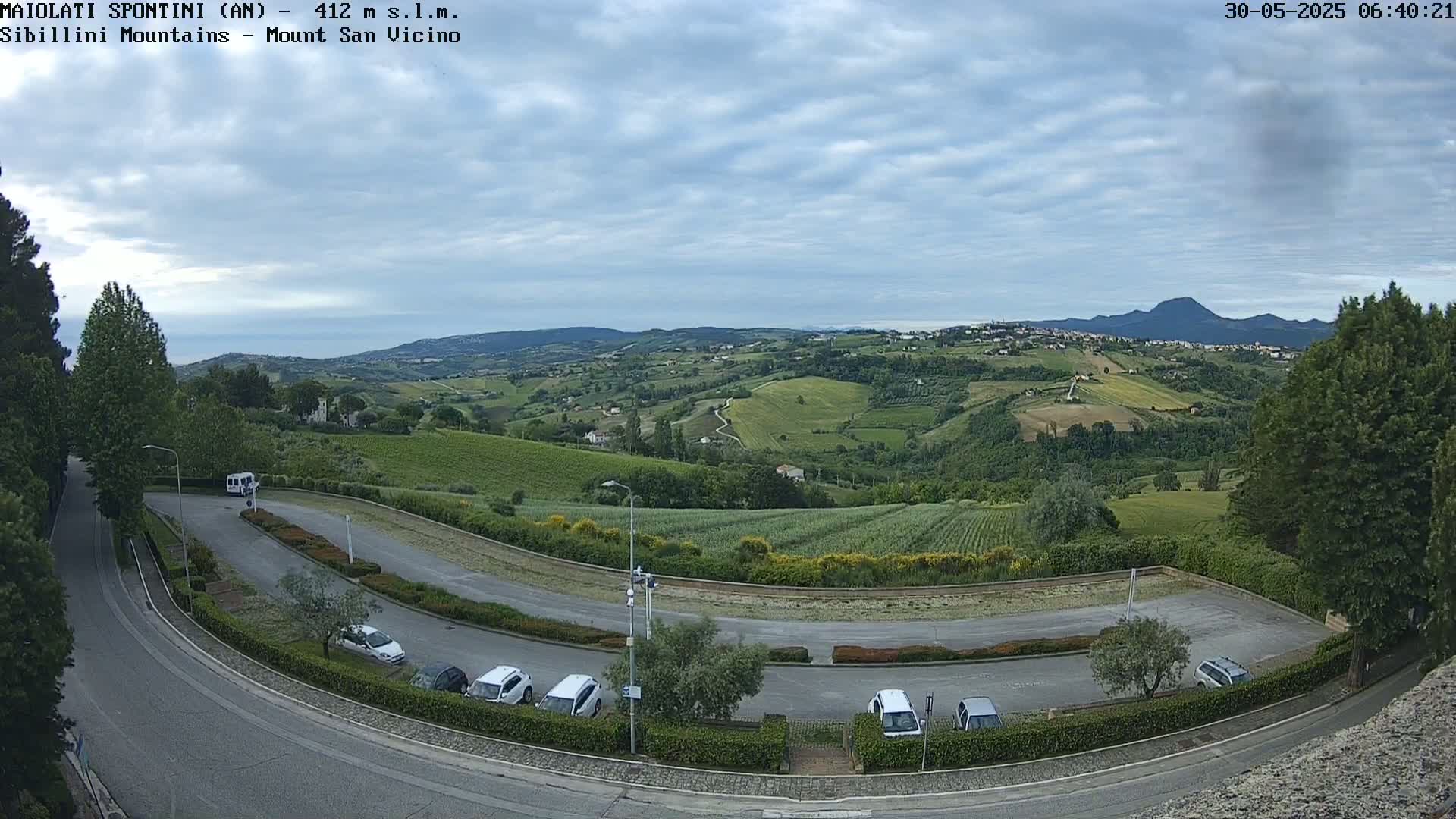 A mostly sunny, partly cloudy day overlooks a hilly landscape with vineyards and a parking lot containing several cars, curving roads, and distant mountains.