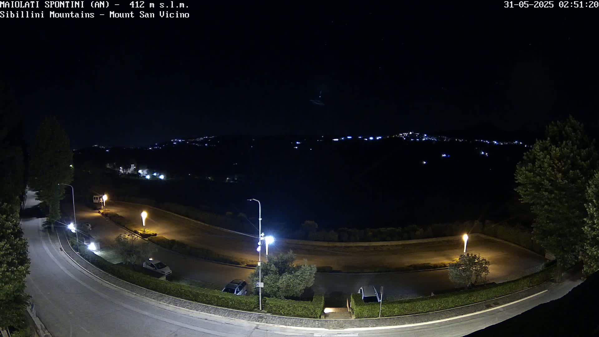A nighttime view of a curving road with parked cars and illuminated lights, overlooking a distant town under a clear, starry sky.