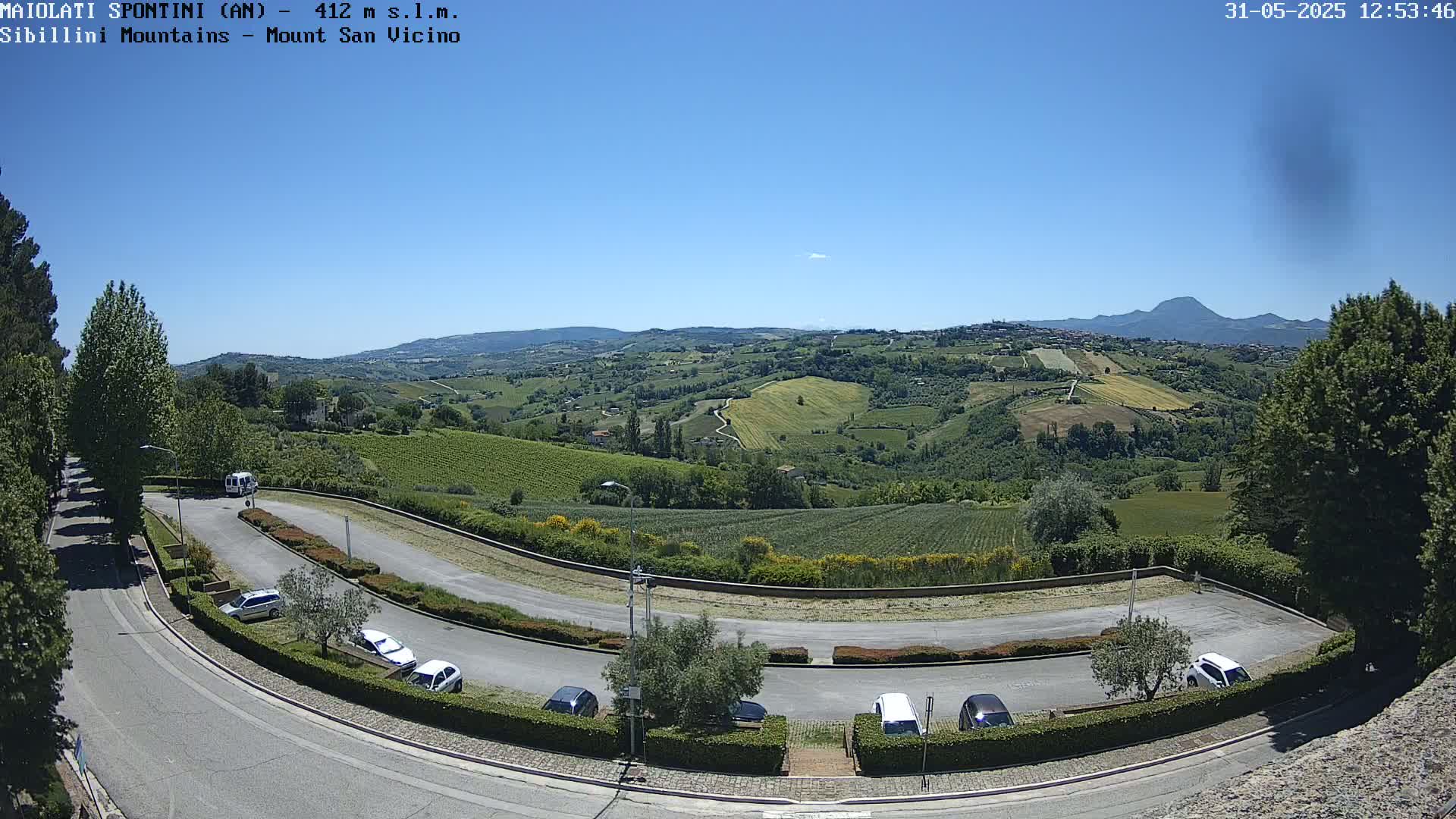 A sunny, clear day overlooking a hilly landscape of vineyards and farmland, with a parking lot and road curving through the foreground.