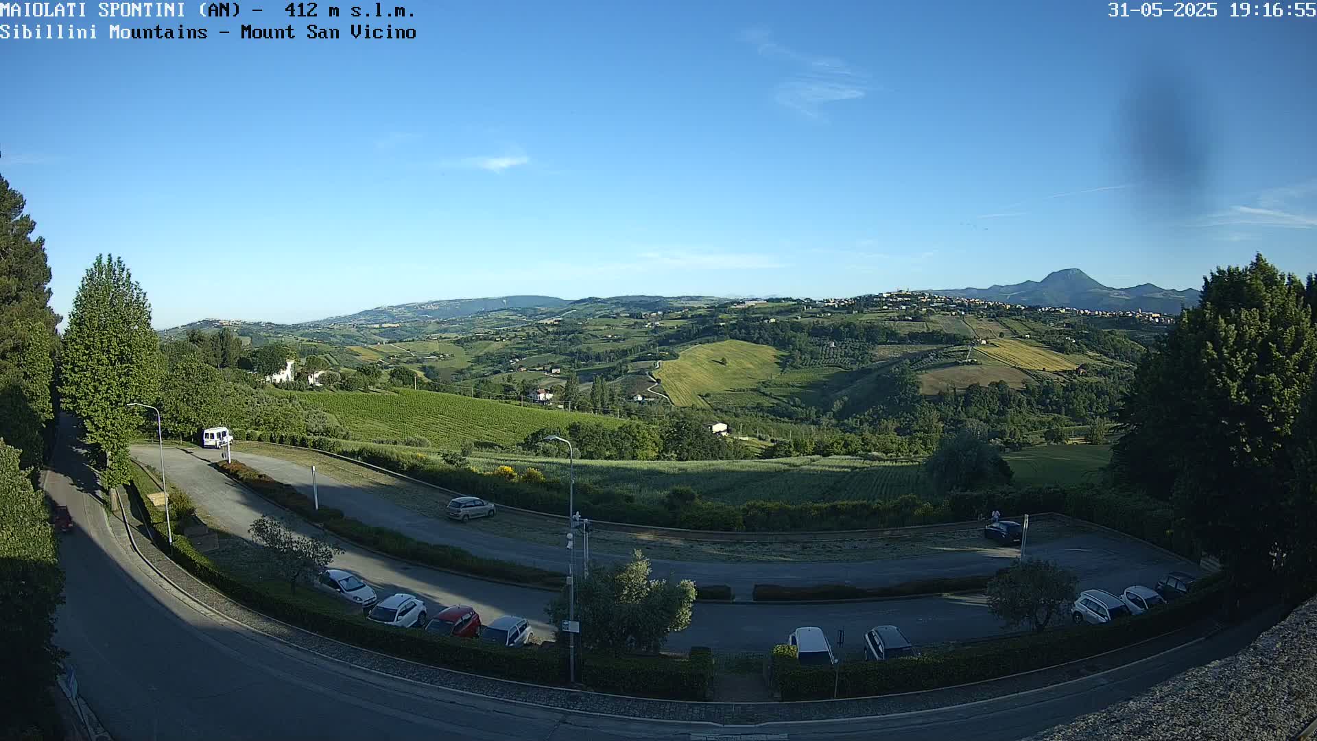 A sunny, clear day overlooking a parking lot and rolling green hills with vineyards, culminating in distant mountains.