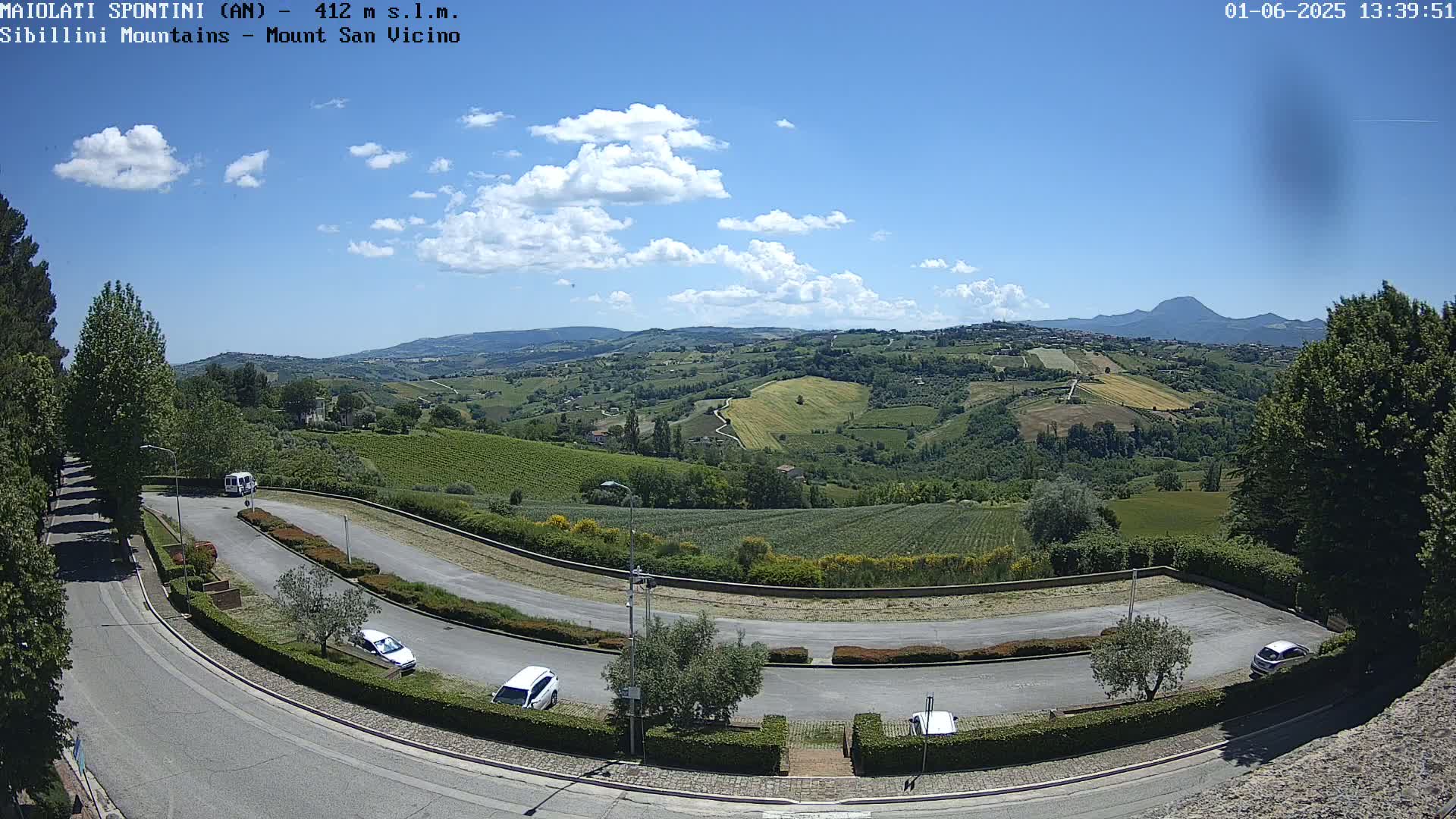 A sunny day reveals a winding road with parked cars overlooking a hilly landscape of vineyards and fields, under a partly cloudy sky.