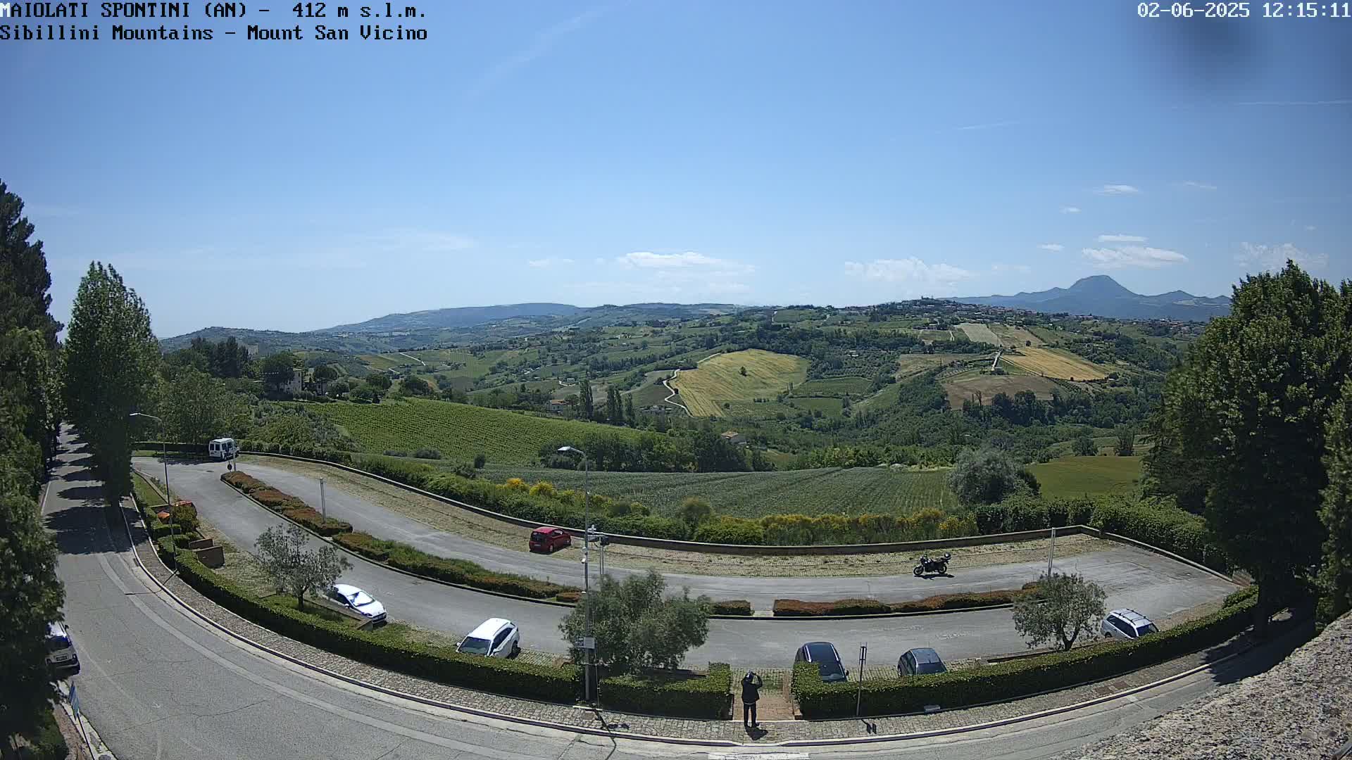 A sunny view of a winding road with parked cars, overlooking rolling hills of vineyards and farmland under a clear blue sky, with a distant mountain range visible in the background.
