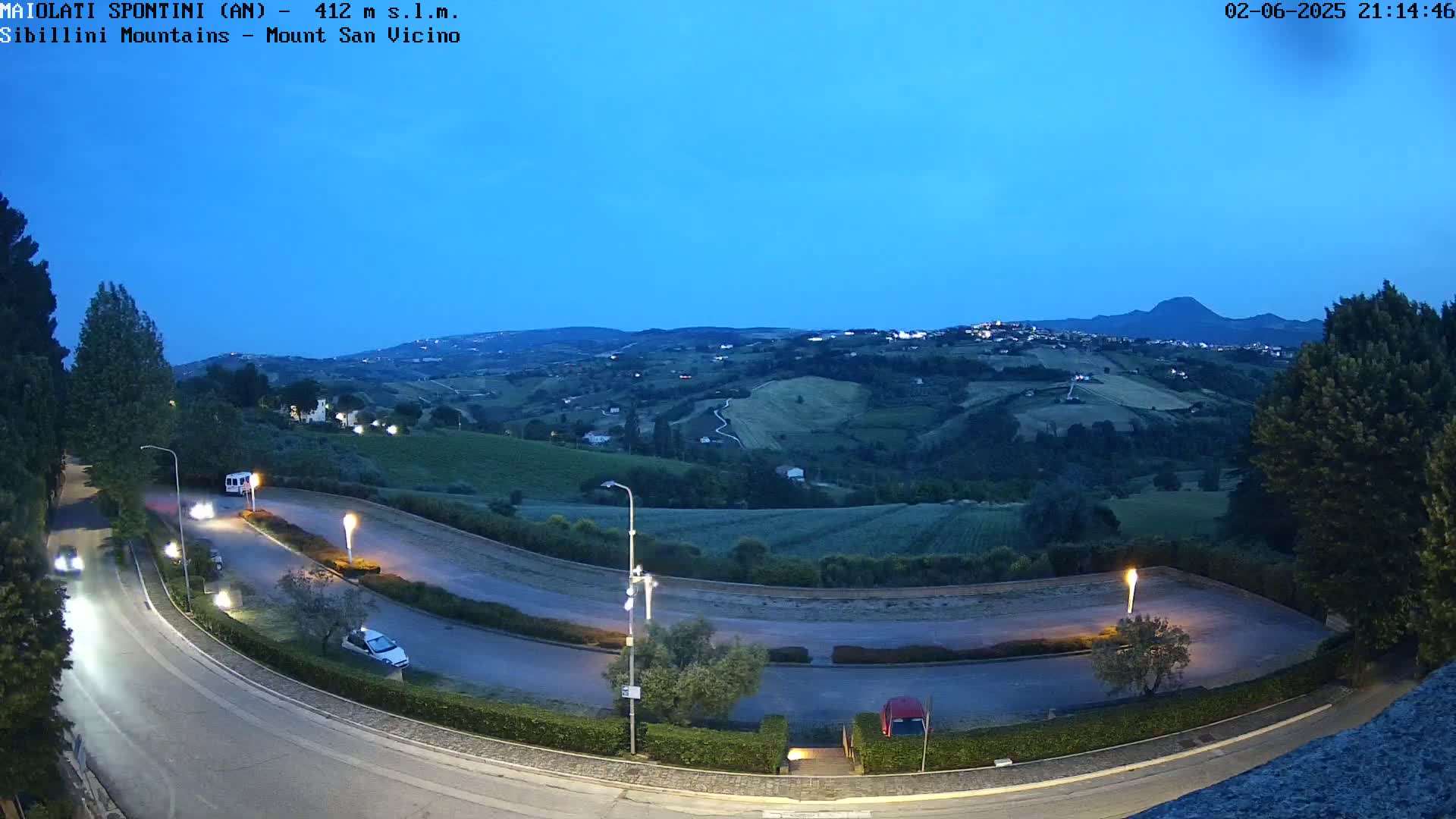 A curved road with several cars and a van is seen at twilight under a clear, dark blue sky overlooking a hilly landscape dotted with lights and greenery.