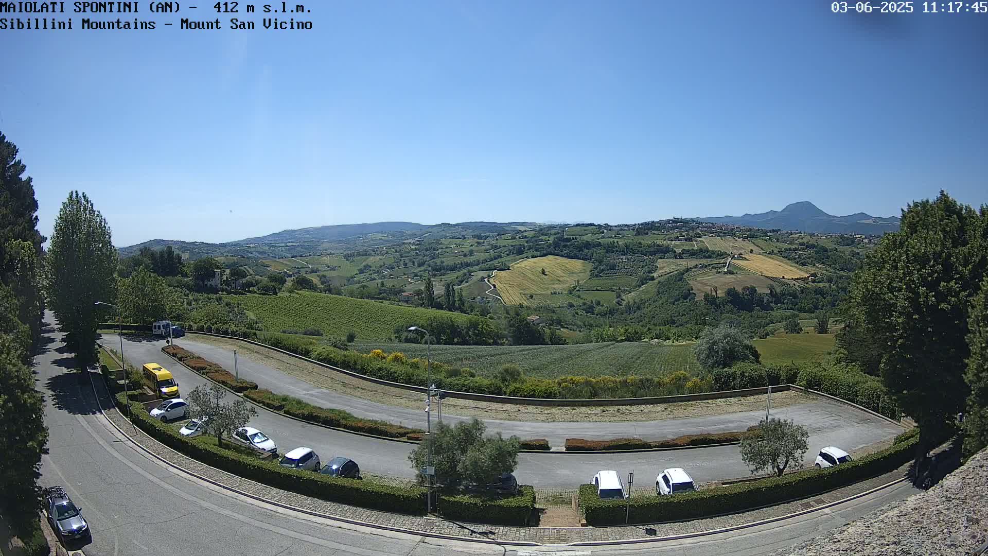 A sunny, clear day reveals a parking lot overlooking a rolling landscape of green hills and vineyards, with a distant mountain range visible in the background.