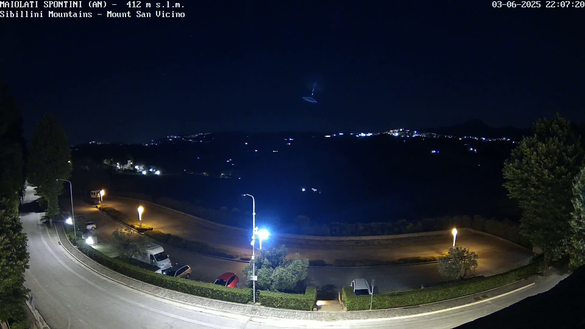 A nighttime, overhead view of a roadway curving through a parking lot and residential area, overlooking a distant town under a clear, starry sky with a light trail visible in the distance.