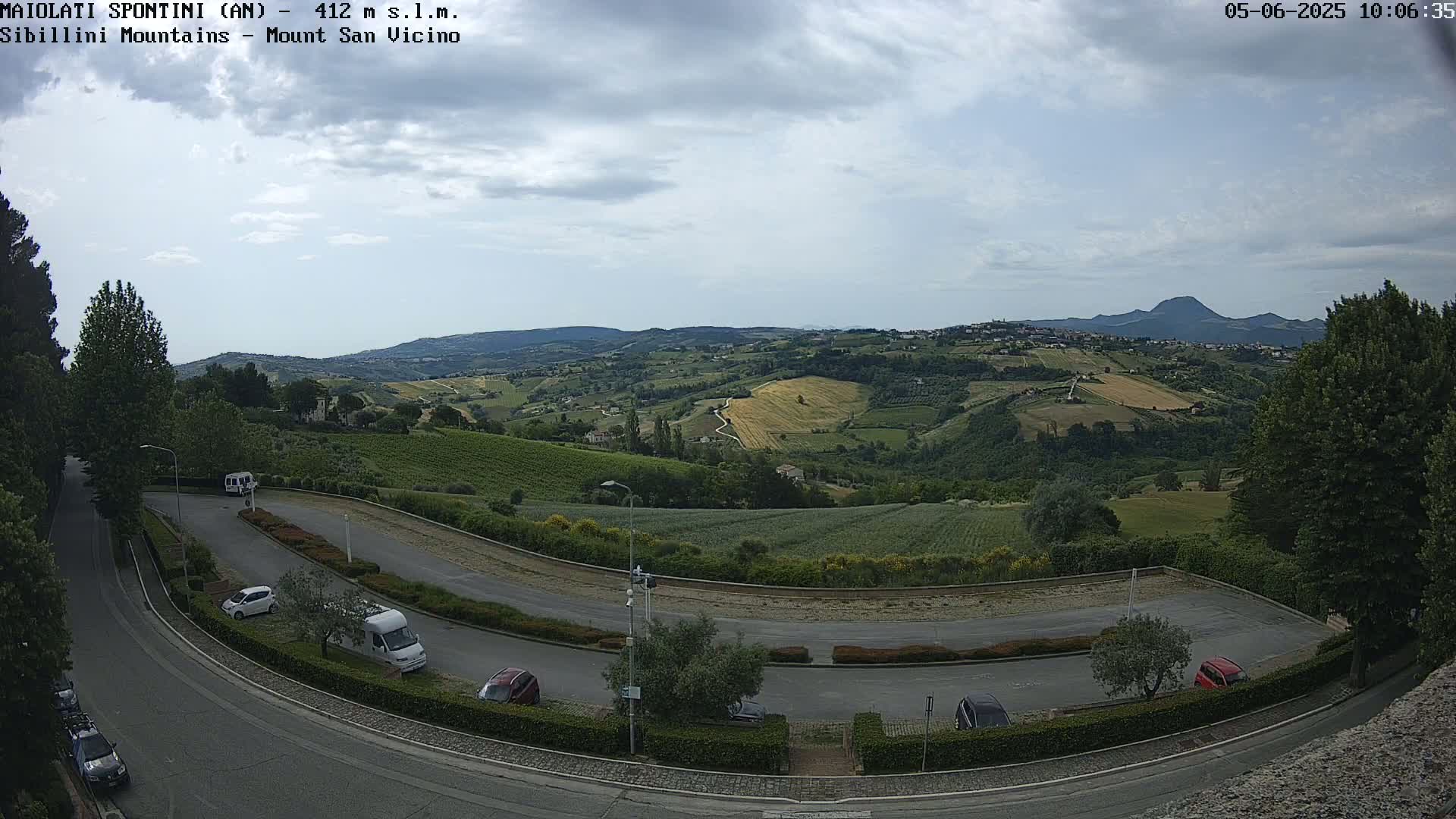 A partly cloudy day overlooks a hilly landscape with vineyards and a parking lot containing several cars and a van.