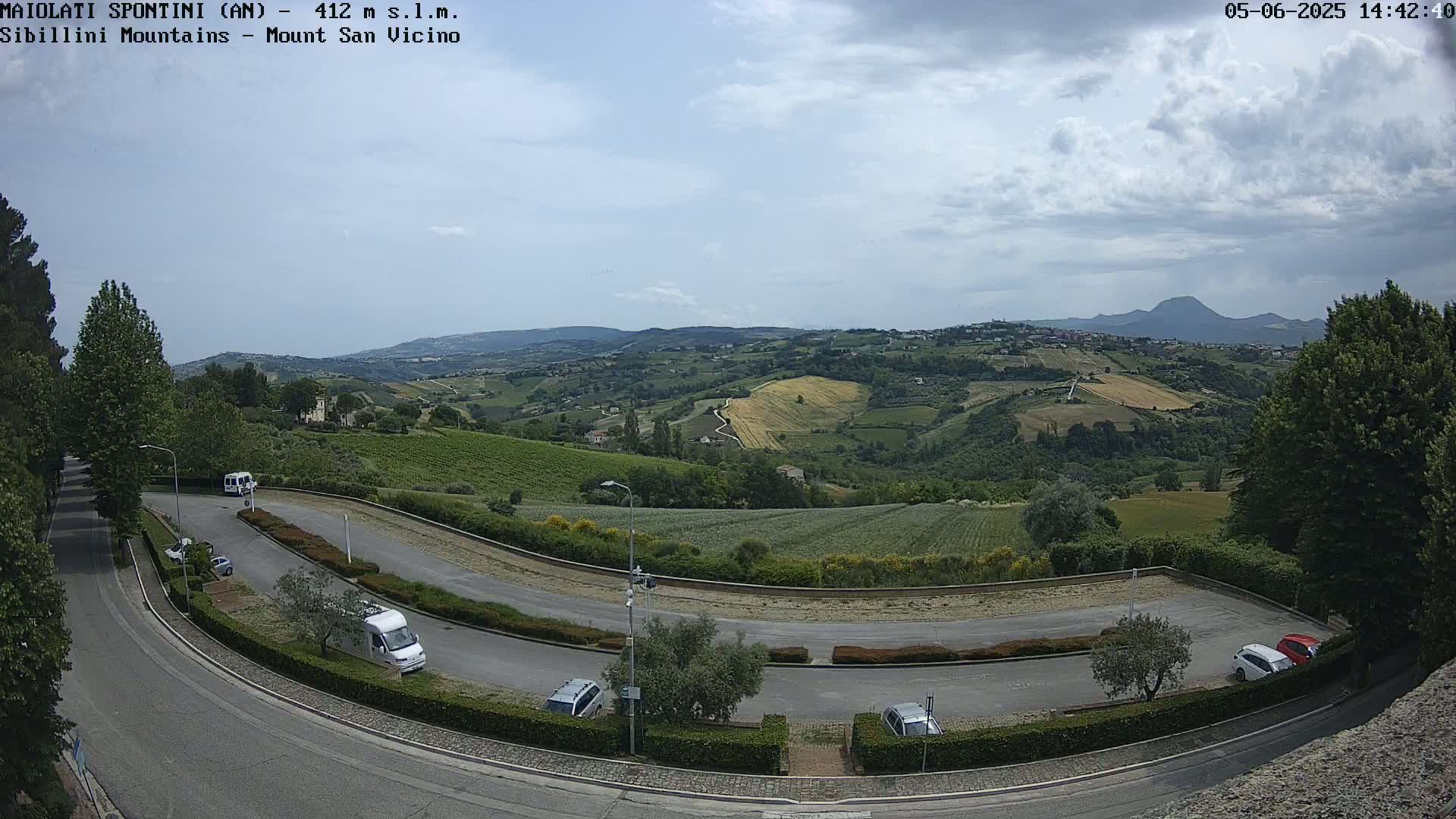 A parking lot with several cars overlooks a gently rolling landscape of vineyards and farmland under a partly cloudy sky.