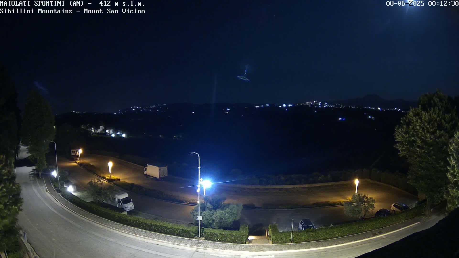 A nighttime view of a road curving through a parking lot with several vehicles, illuminated by streetlights, overlooks a distant town nestled in dark hills under a clear, dark sky.