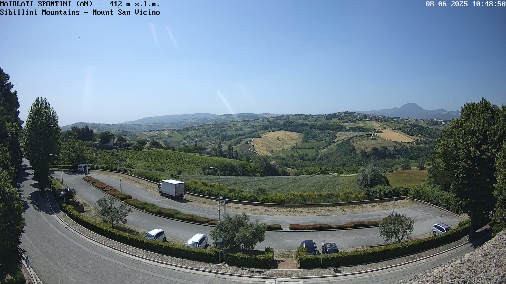 A sunny, expansive view of a hilly landscape with vineyards and a parking lot containing several cars and a truck, overlooked by a curving road.