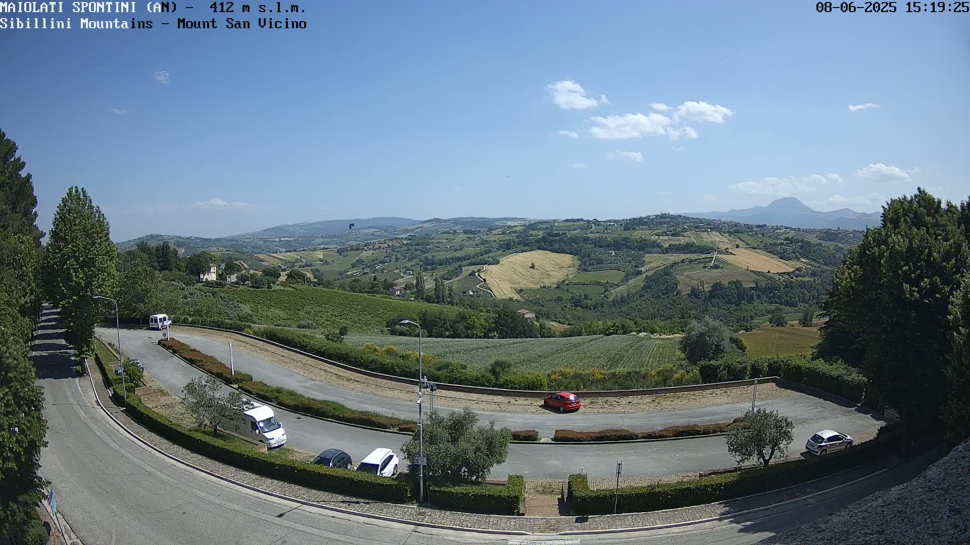 A sunny day reveals a winding road with several parked cars overlooking a picturesque landscape of rolling green hills and distant mountains.