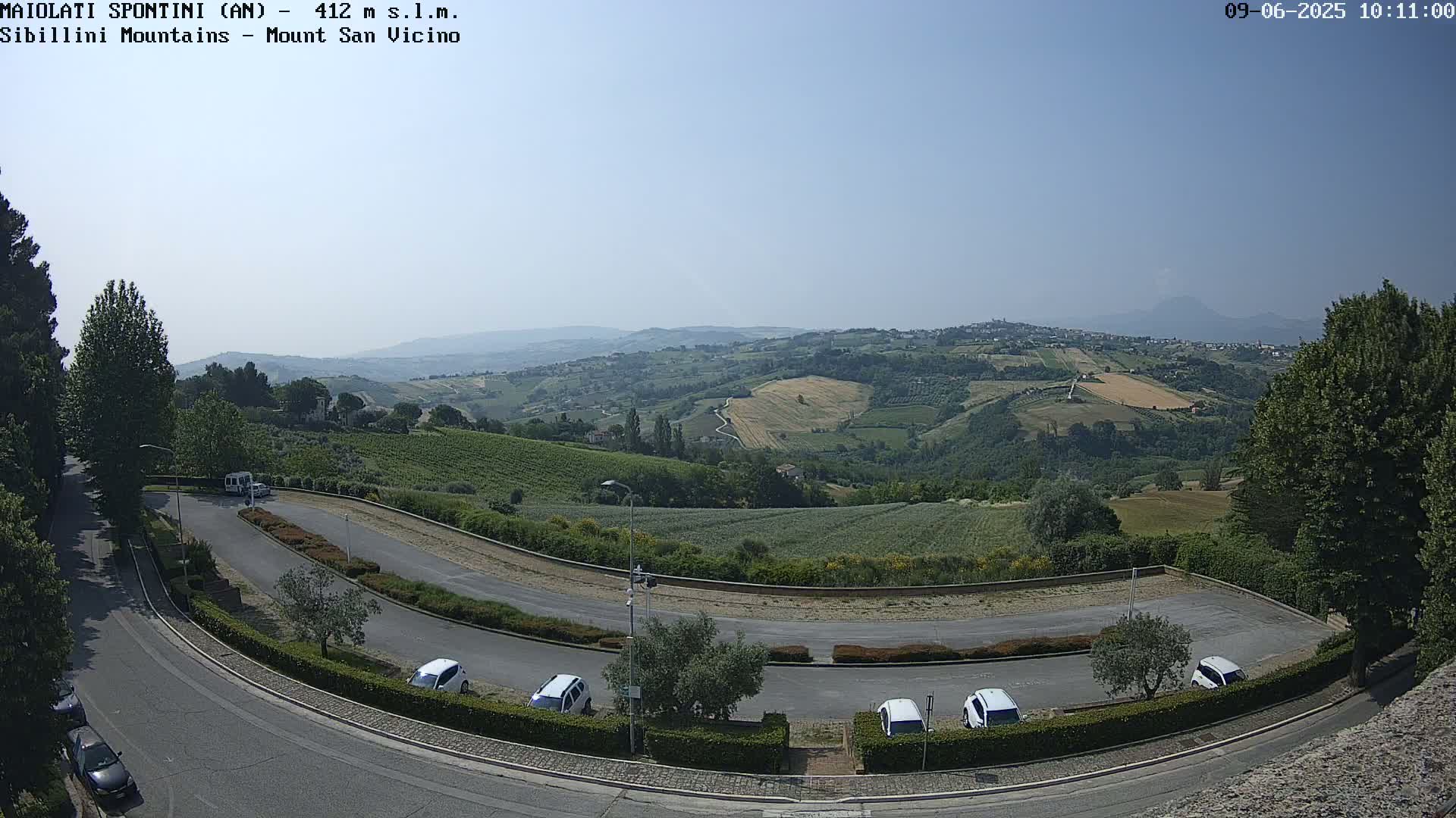 A curved road and parking lot overlook a sunny valley dotted with farms and vineyards under a clear blue sky.