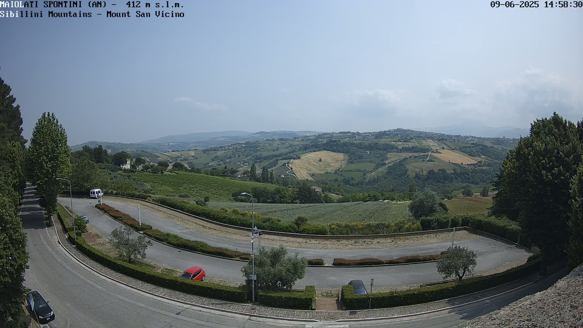 A sunny, high-angle view shows a winding road curving through a parking area and leading to a vista of rolling green hills and farmland under a clear sky.