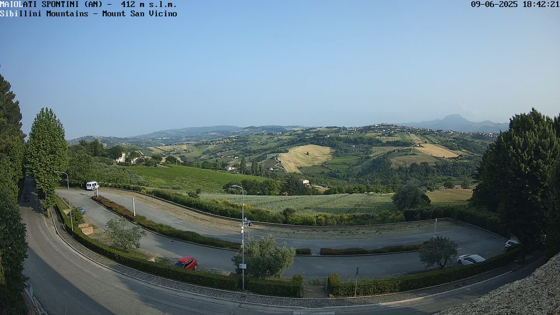 A high-angle, long shot shows a sunny, clear-day view of a winding road curving through a parking lot and leading toward rolling hills and distant mountains.