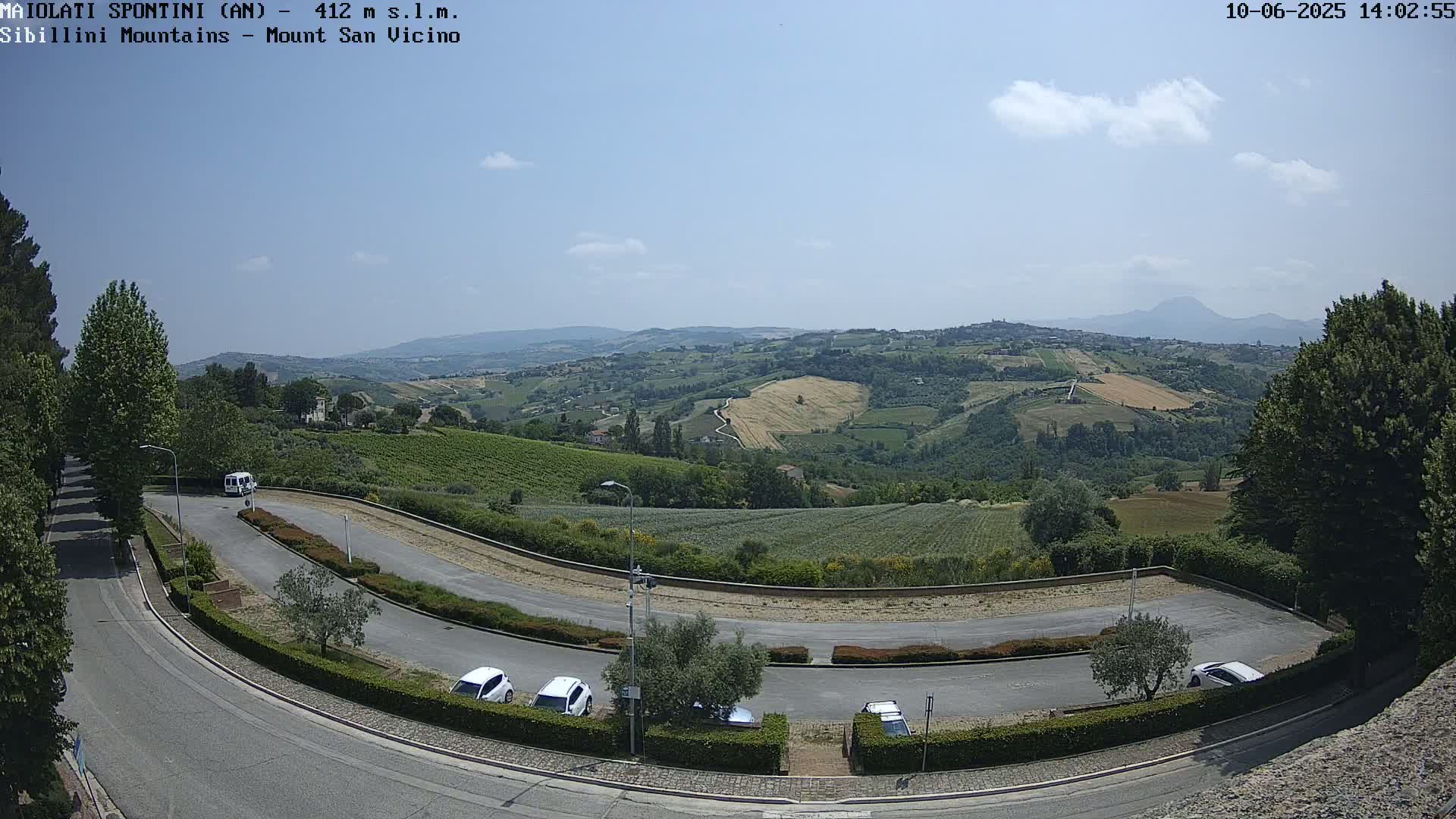 A curved road with parked cars overlooks a sunny, hilly landscape dotted with fields and vineyards under a mostly clear sky.