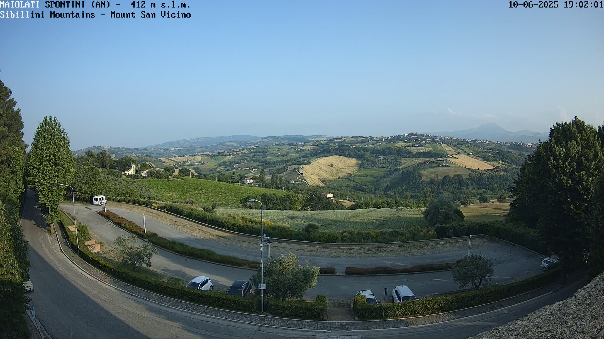 A paved roadway curves through a parking lot overlooking a sun-drenched valley of rolling hills and farmland under a clear blue sky.