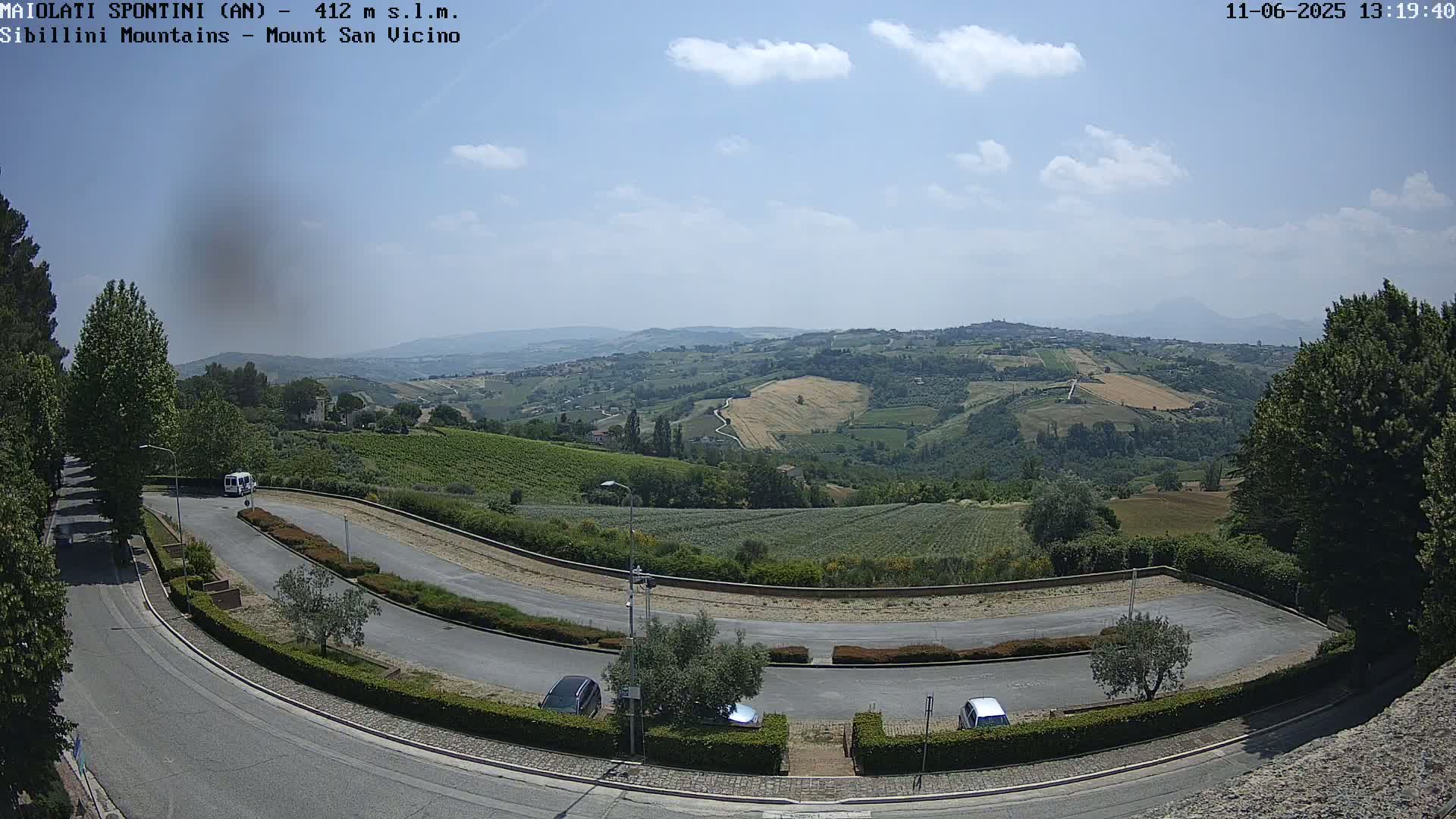 A sunny, partly cloudy day overlooks a winding road and parking area nestled among rolling hills and farmland, with mountains visible in the distance.