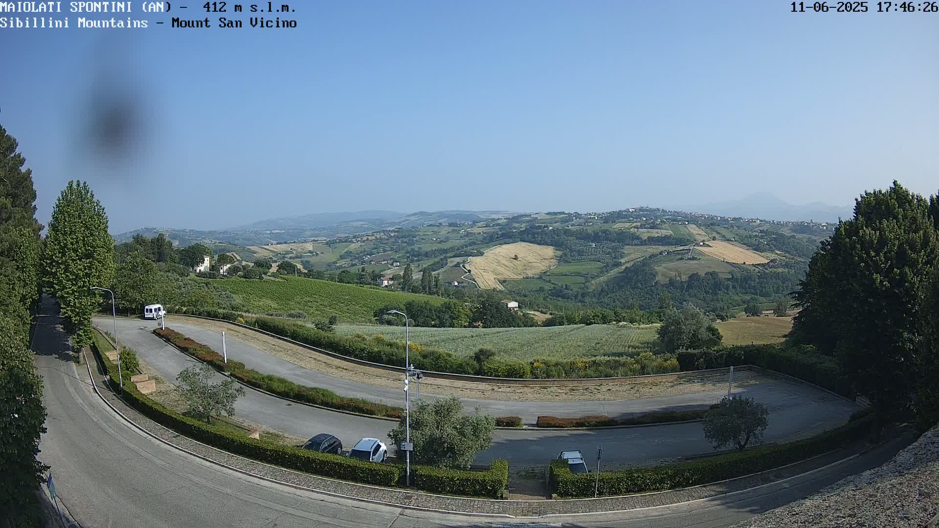 A panoramic view on a clear, sunny day shows a winding road curving through a parking lot with several cars, leading toward rolling hills and fields of varied crops under a bright blue sky.