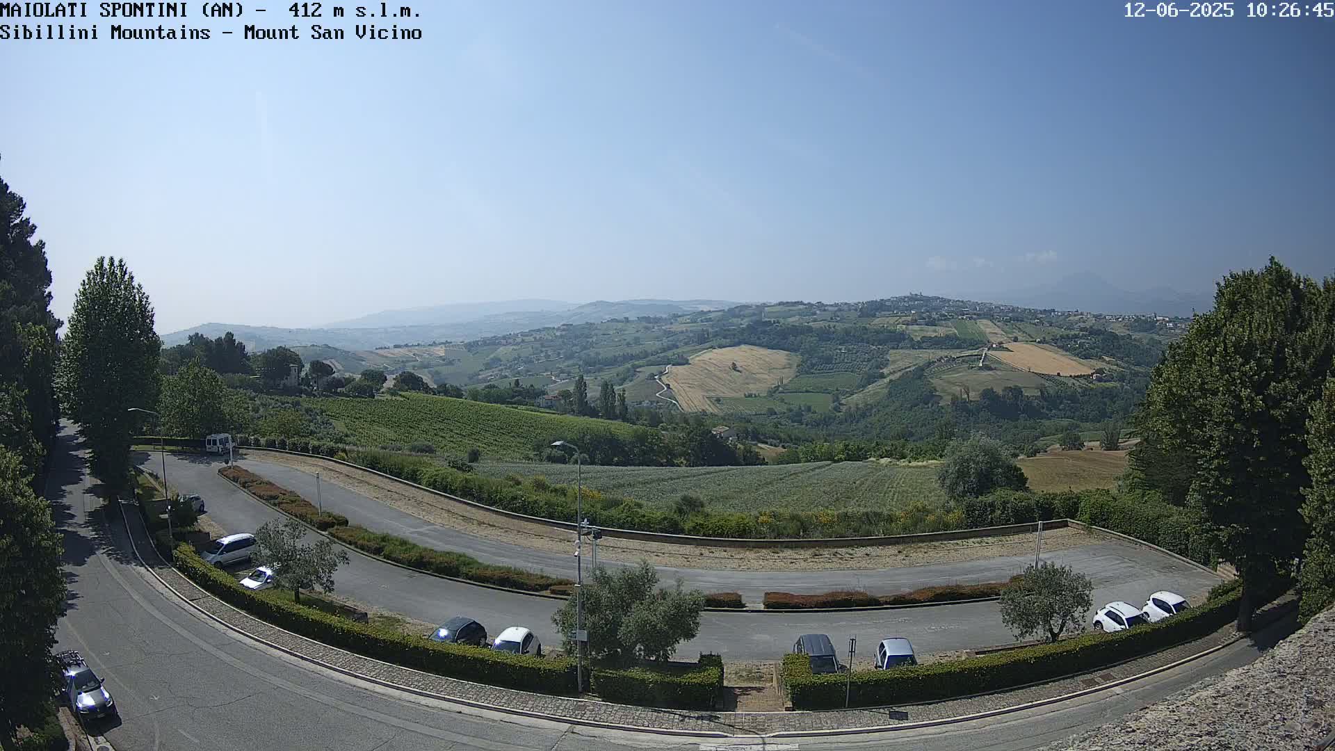 A curving road with parked cars leads to a panoramic view of rolling hills and distant mountains under a clear, sunny sky.