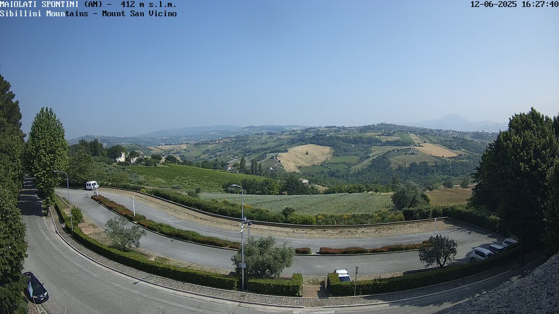 A winding road curves through a parking lot overlooking a sunny, rolling landscape of green hills and fields under a clear blue sky.
