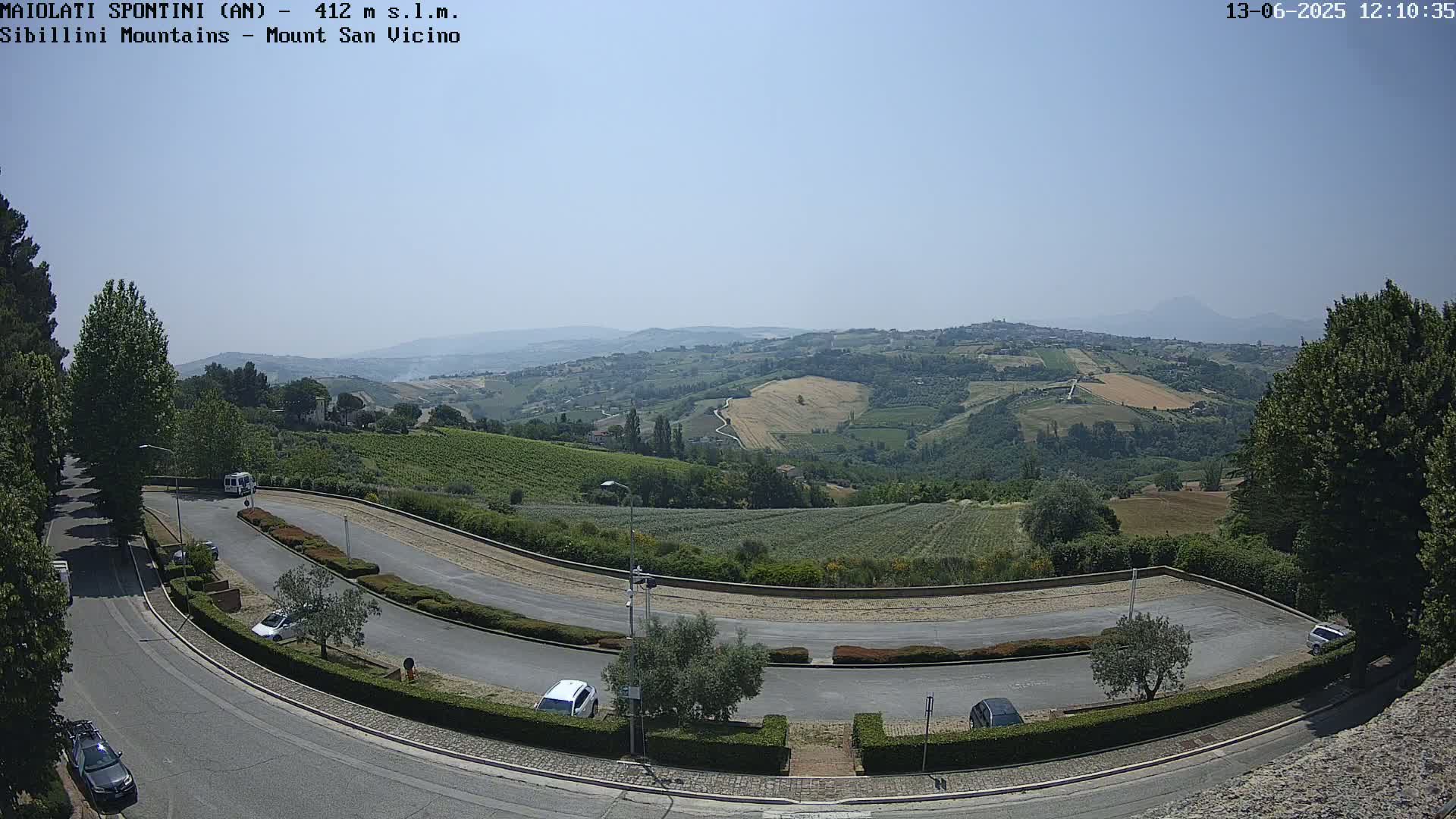 A winding road curves through a parking lot overlooking a sun-drenched valley of vineyards and farmland, with distant mountains visible under a clear sky.