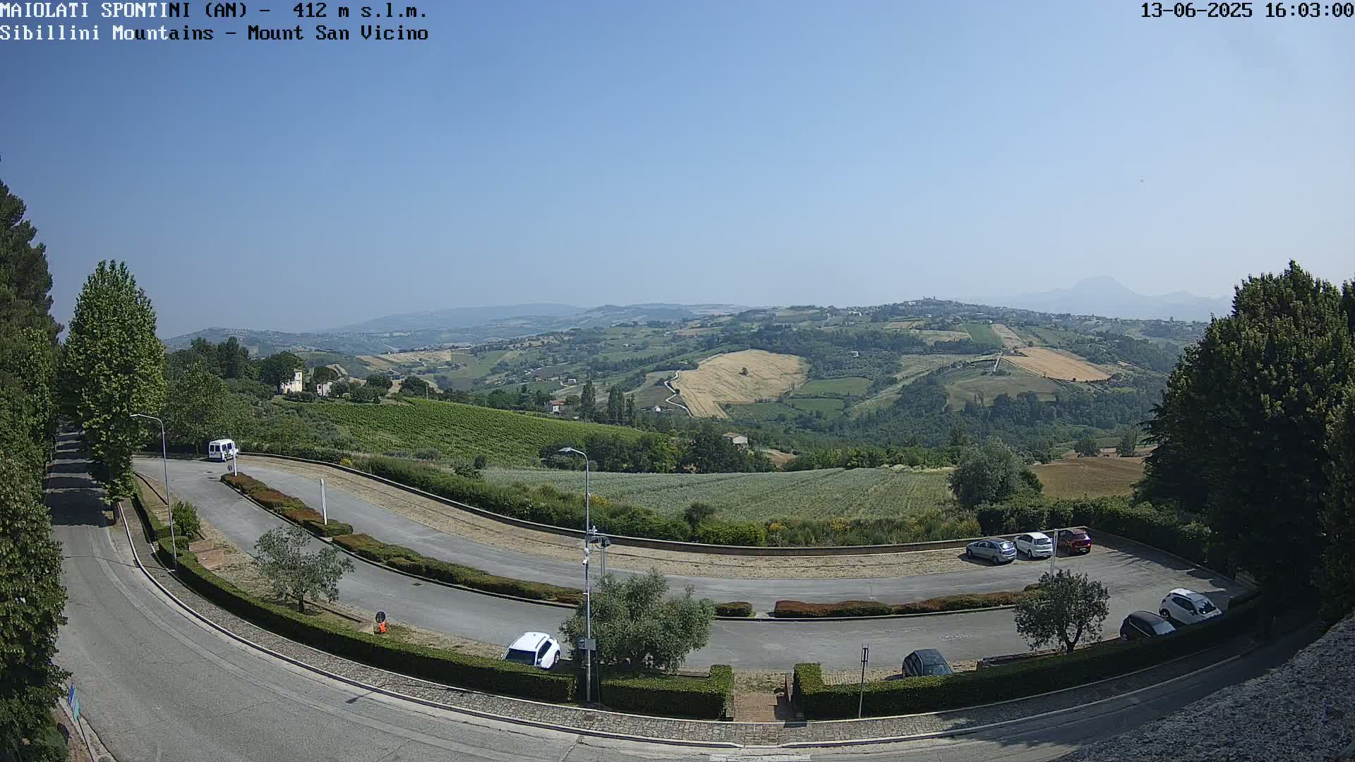 A winding road curves through a parking lot overlooking a sun-drenched valley of rolling hills and fields under a clear blue sky.