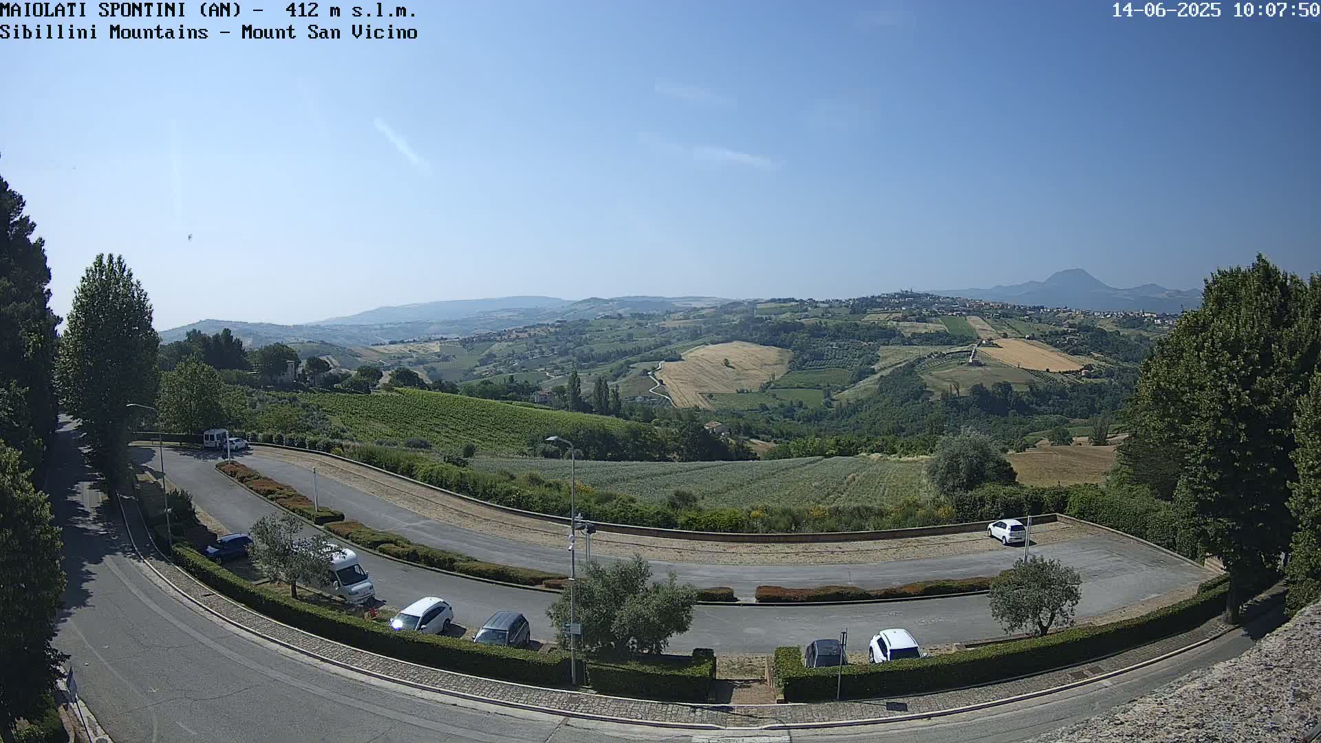 A sunny, clear day overlooks a winding road and parking lot nestled within a hilly landscape dotted with fields and vineyards, with distant mountains visible on the horizon.