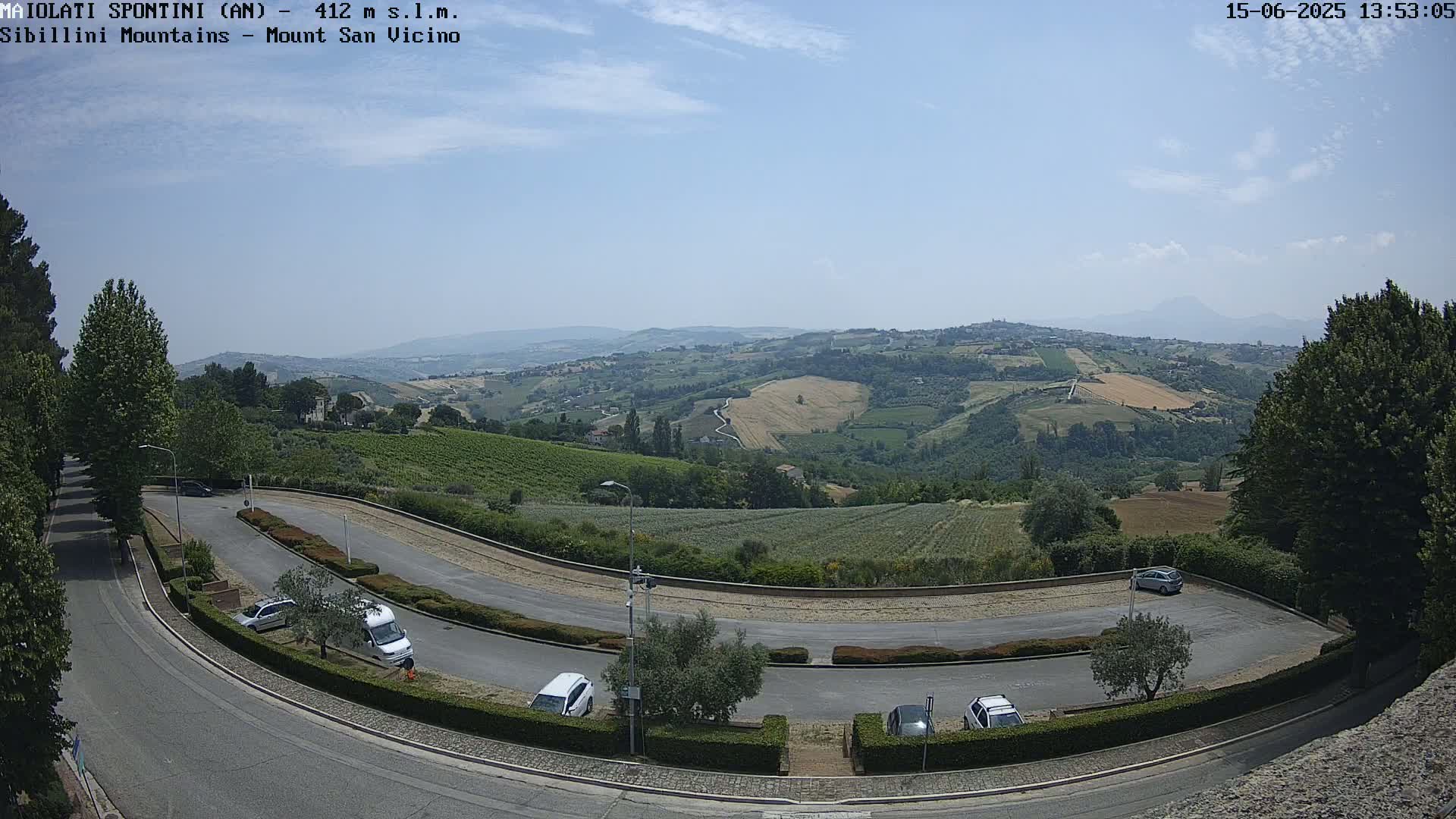 A curving road with parked cars overlooks a sunny, hilly landscape dotted with fields and vineyards under a mostly clear sky.