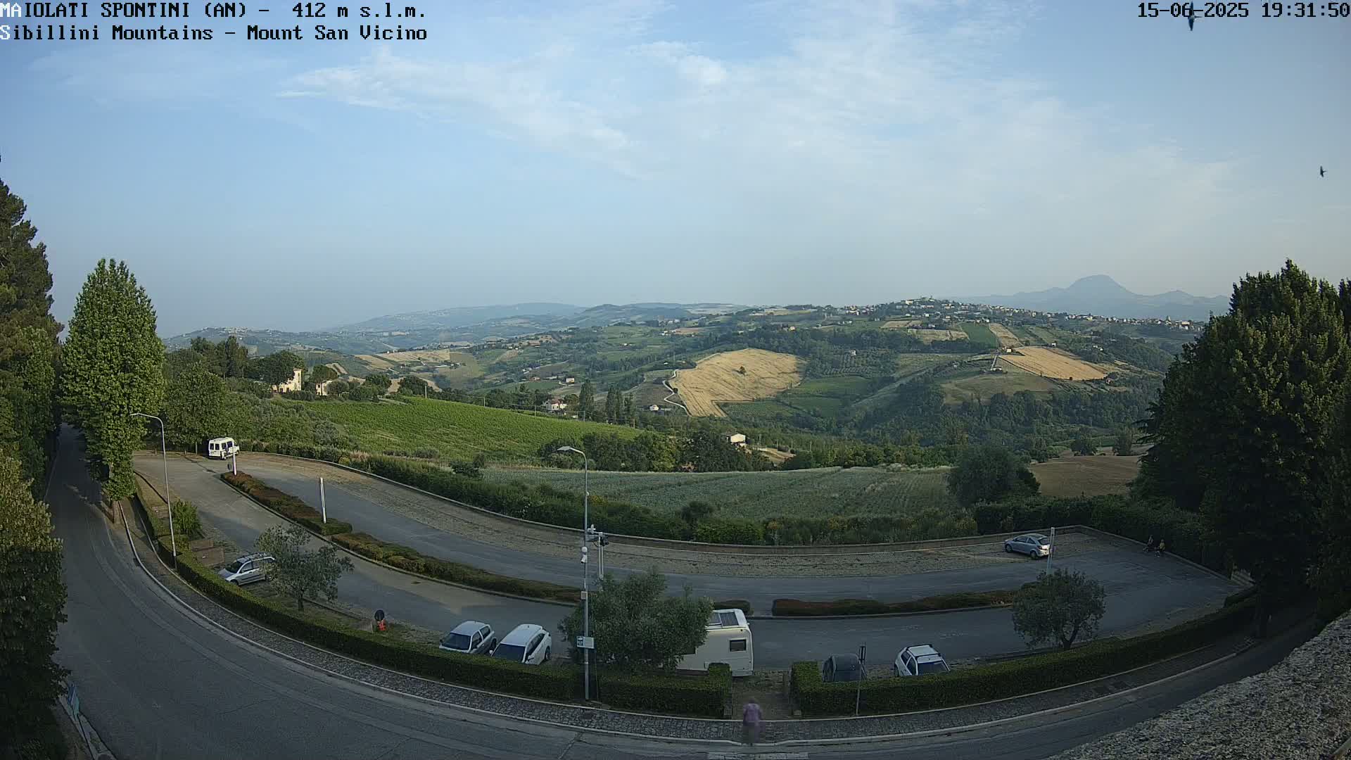 A high-angle view shows a parking lot and winding road overlooking a hilly landscape of vineyards and farmland under a mostly clear sky.