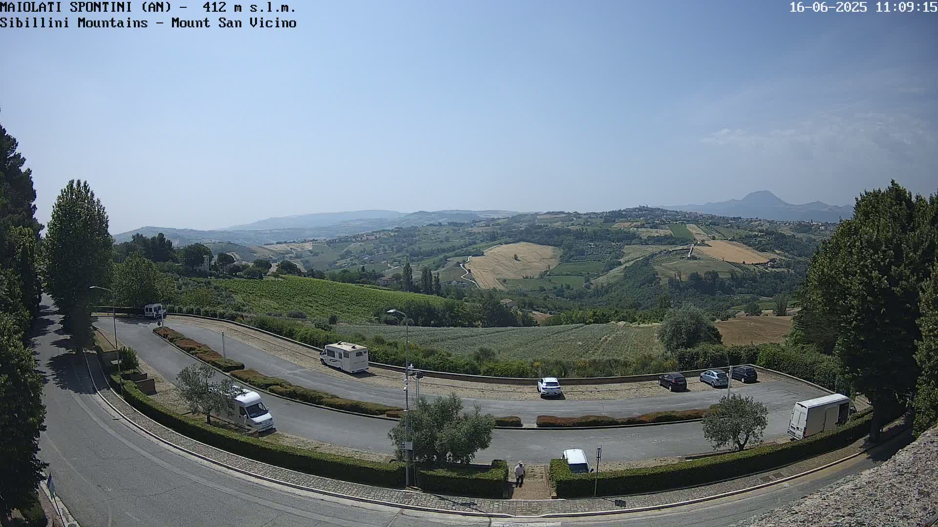 A sunny, clear day overlooks a winding road with parked cars and recreational vehicles, situated in a hilly landscape with vineyards and distant mountains.
