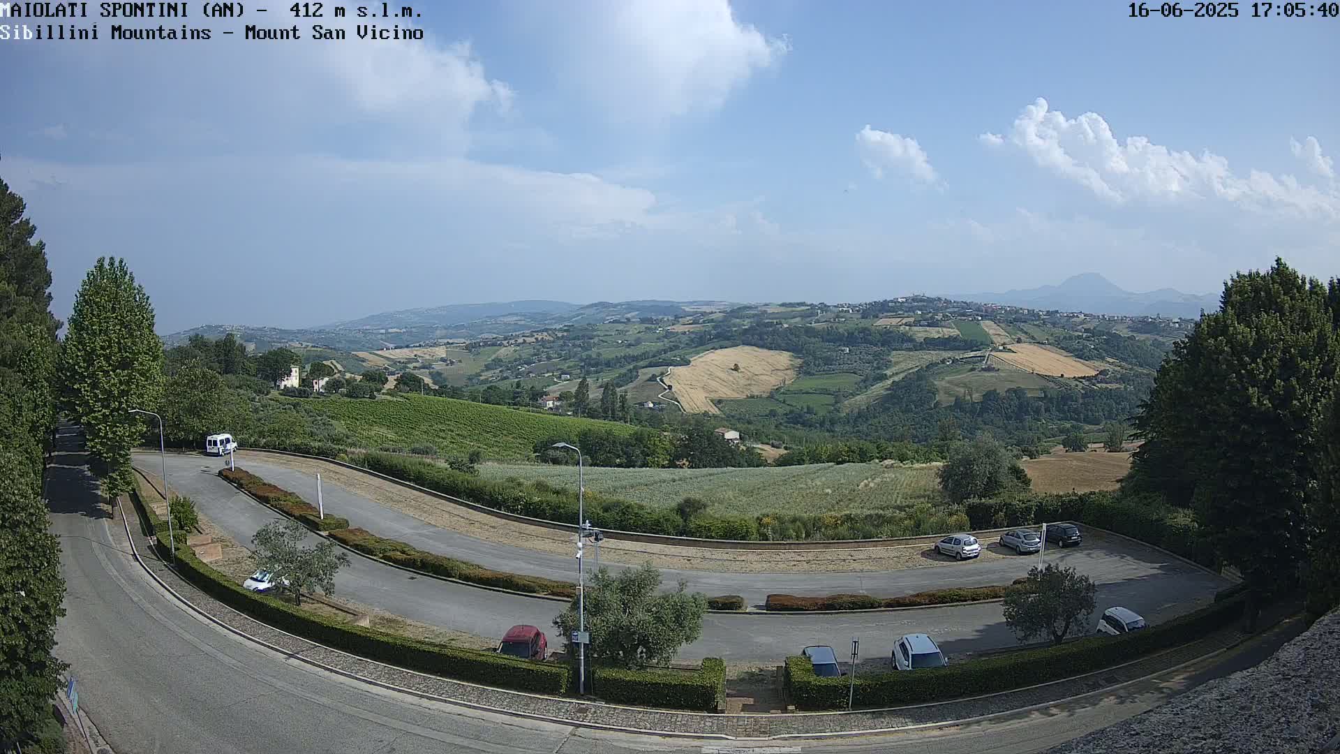A partly cloudy summer day overlooks a winding road and parking lot nestled among rolling green hills and fields, with distant mountains visible in the background.