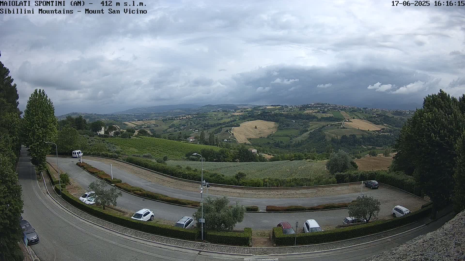 A hillside parking lot with several cars overlooks a valley of rolling green hills under a partly cloudy sky.